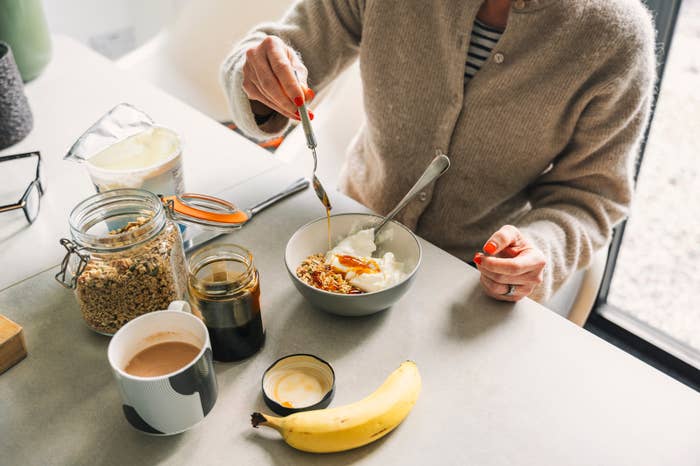 Person preparing yogurt with granola astatine  a table, surrounded by coffee, banana, and jars