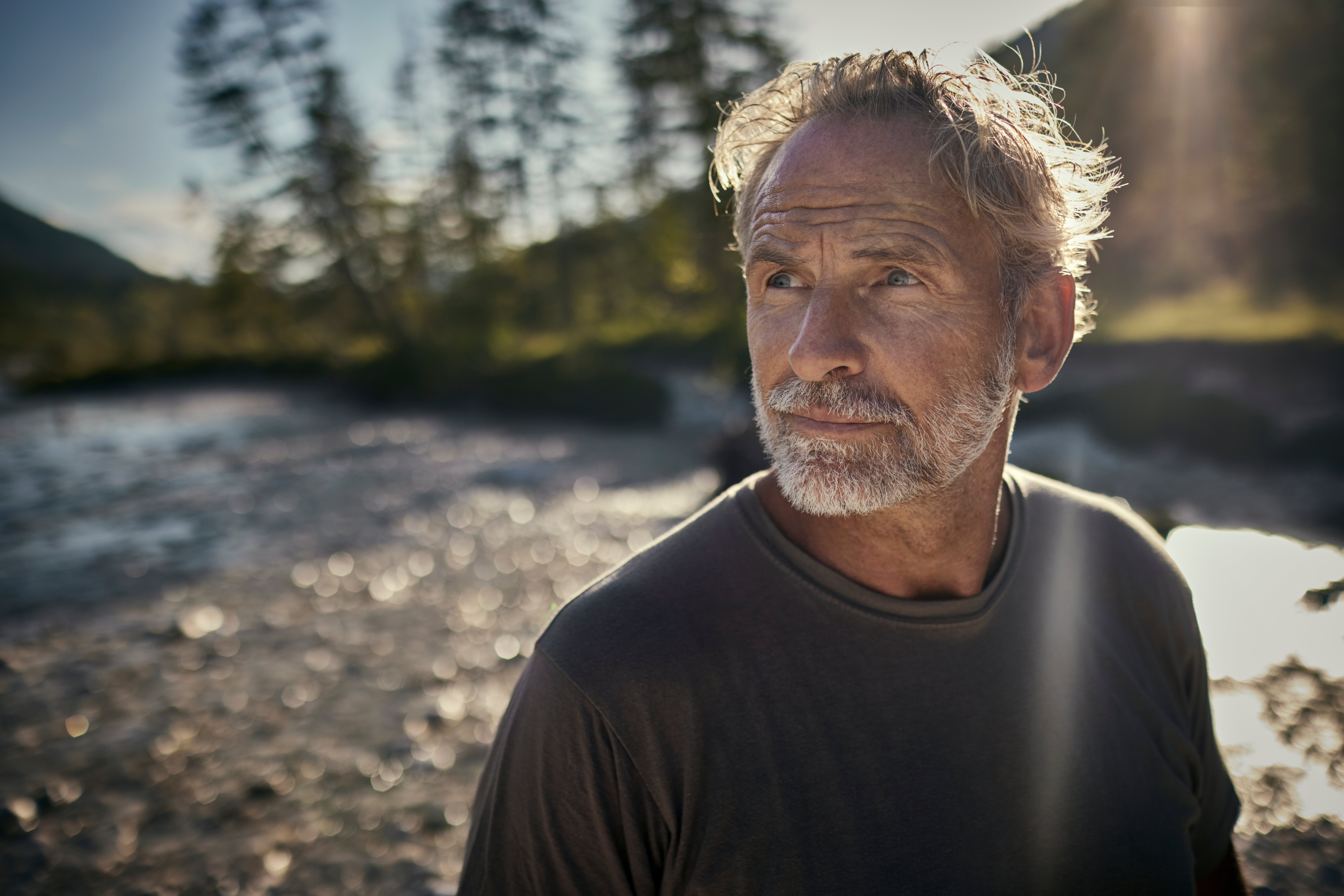 A thoughtful antheral with grey hairsbreadth and a beard stands outdoors adjacent a river, looking into the distance. The country suggests contemplation and well-being