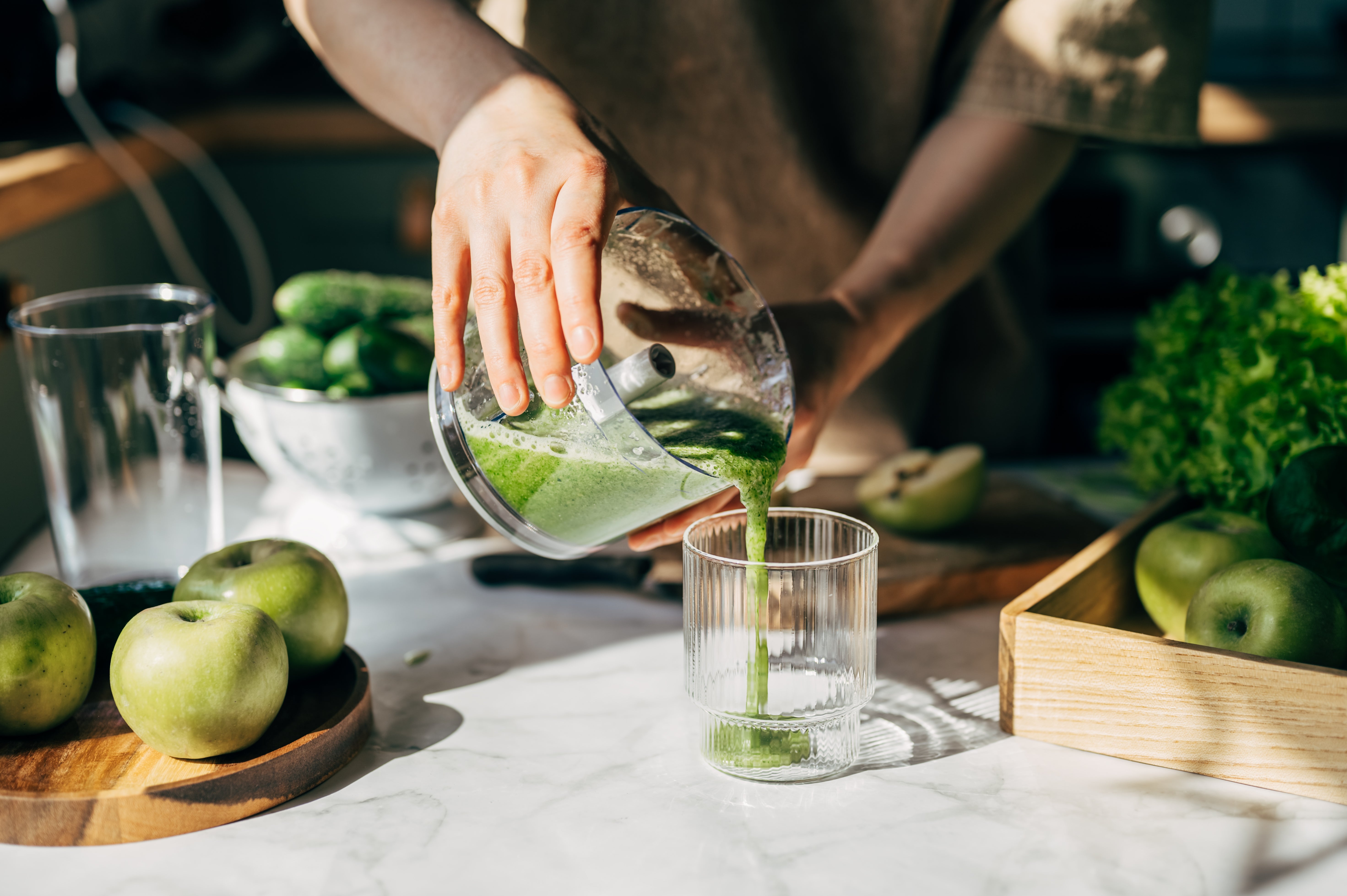 Person pouring greenish smoothie into a glass; surrounded by caller apples, cucumbers, and lettuce, suggesting a absorption connected steadfast living