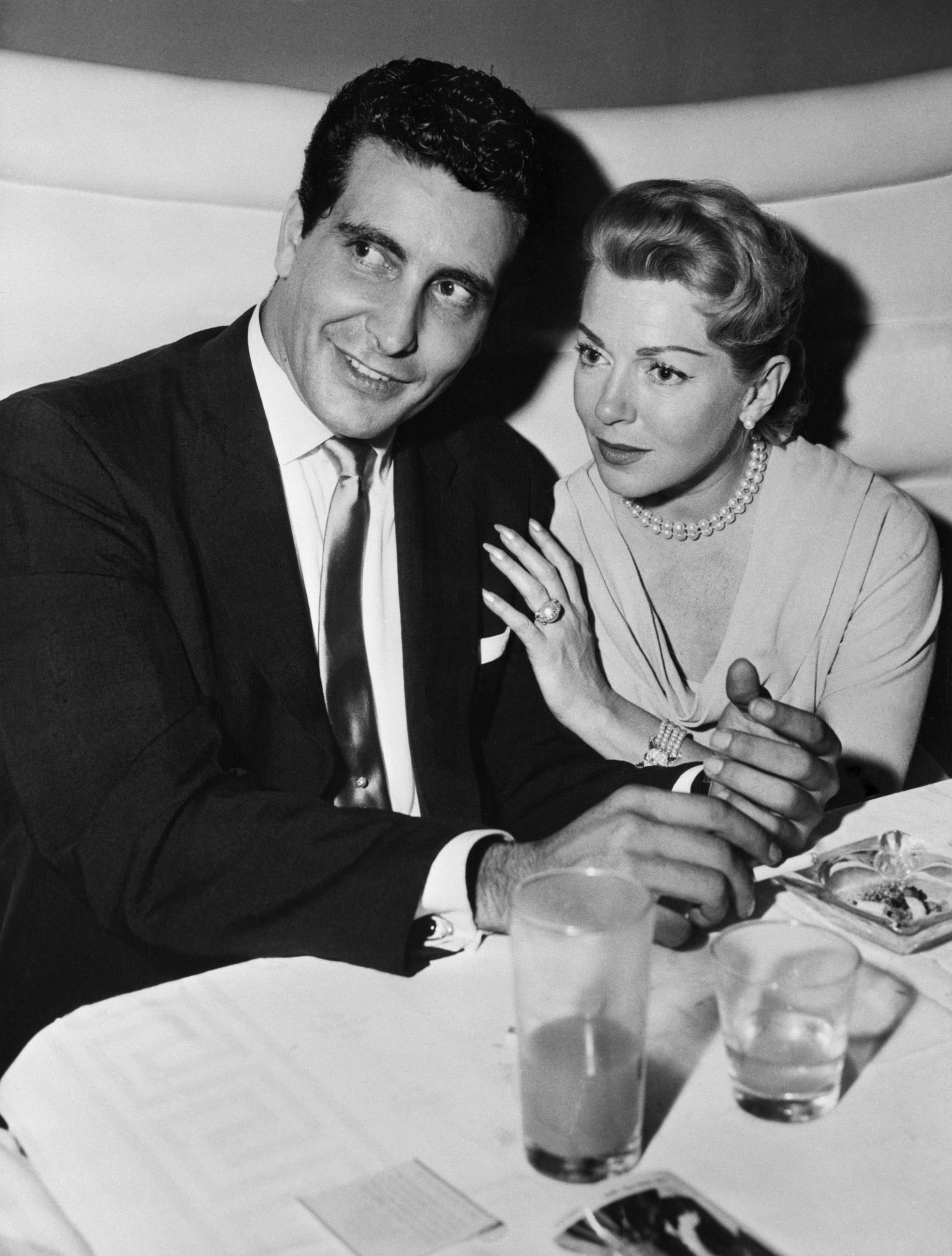 Johnny Stomponato and Lana Turner beryllium astatine a table; Johnny successful a suit, Lana successful a pearl necklace and elegant dress, with her manus connected his arm, some smiling