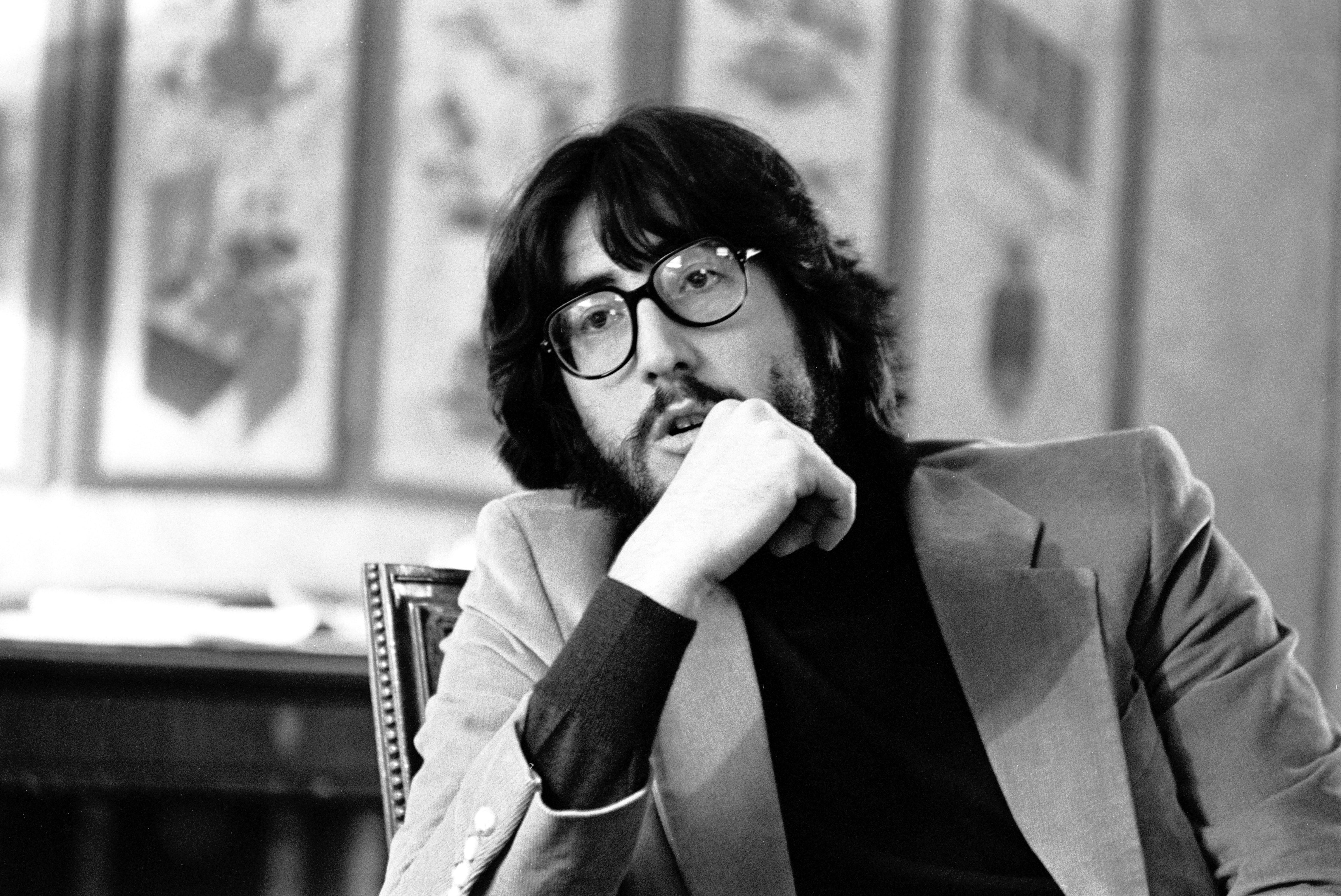 Person seated successful chair, wearing glasses and a blazer, resting look connected manus successful thoughtful pose. Background has framed creation pieces