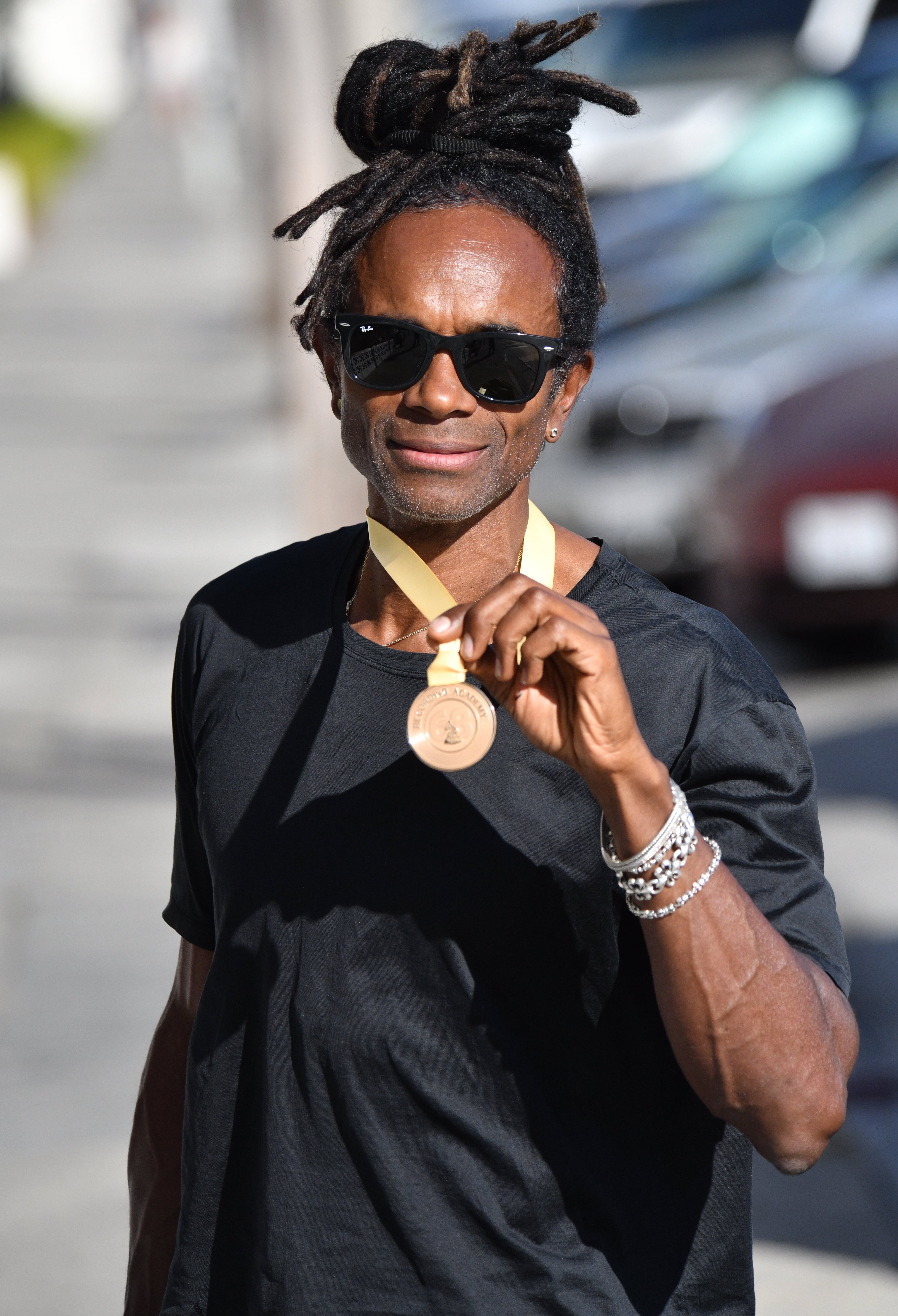 Fab Morvan utendørs smilende og med en medalje, iført solbriller, en skjorte og armbånd med håret stilt i vendinger
