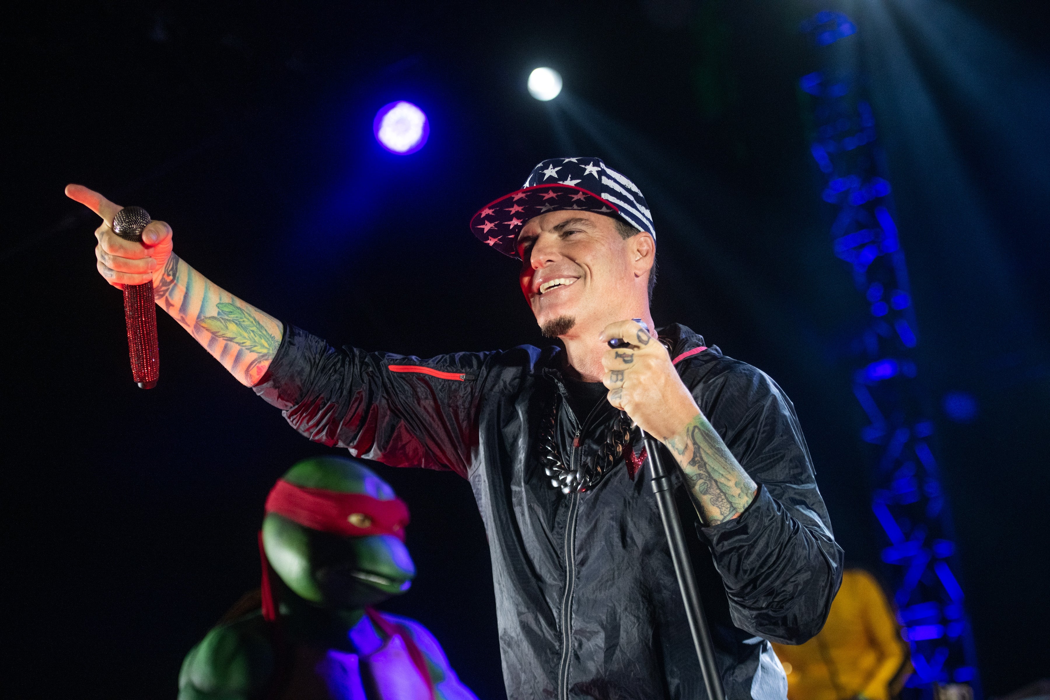Vanilla Ice i en stjernemønstret caps og jakkepeker med et smil på scenen, med en kostymefigur i bakgrunnen