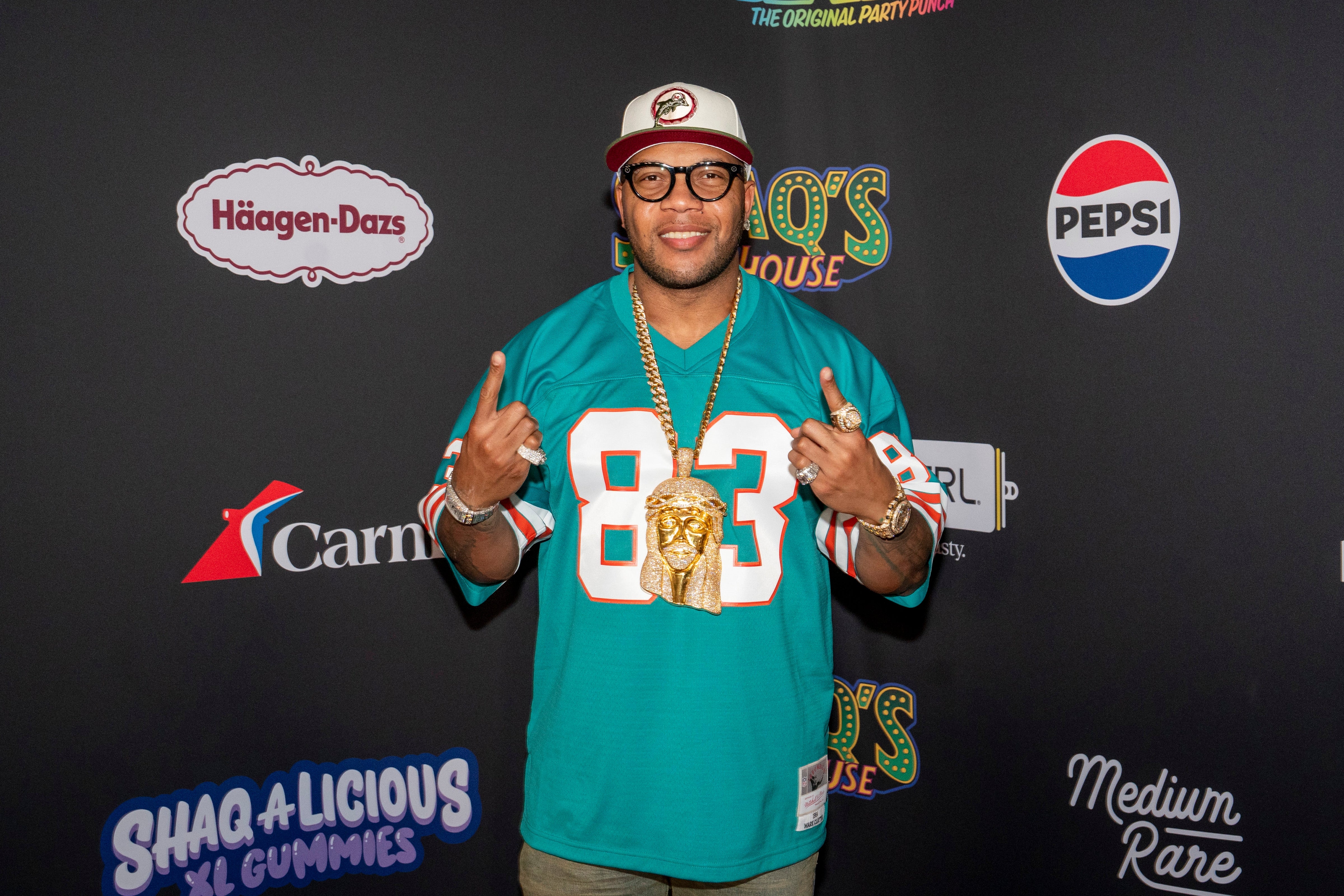 Flo Rida i sportstrøye og caps poserer på et merkede bakteppe på et kjendisarrangement