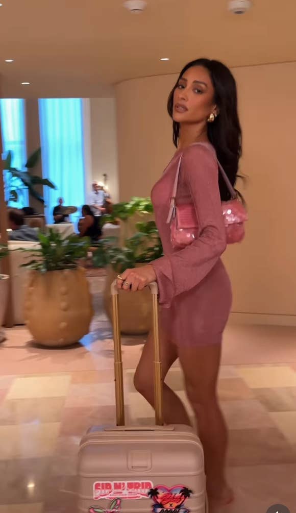 Shay Mitchell in a stylish, form-fitting dress walks with a suitcase in a hotel lobby