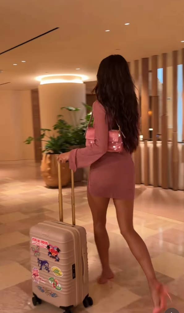 Shay Mitchell in a stylish, form-fitting mini dress, walking barefoot in a hotel lobby, pulling a suitcase adorned with colorful stickers