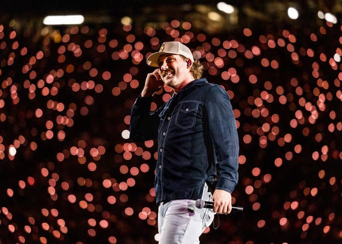 Morgan Wallen connected  signifier    holding a microphone, wearing a casual overgarment   and cap, smiles with agleam  lights successful  the background