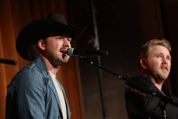 Two musicians perform on stage, one wearing a cowboy hat and denim jacket, the other in a dark jacket, each holding microphones