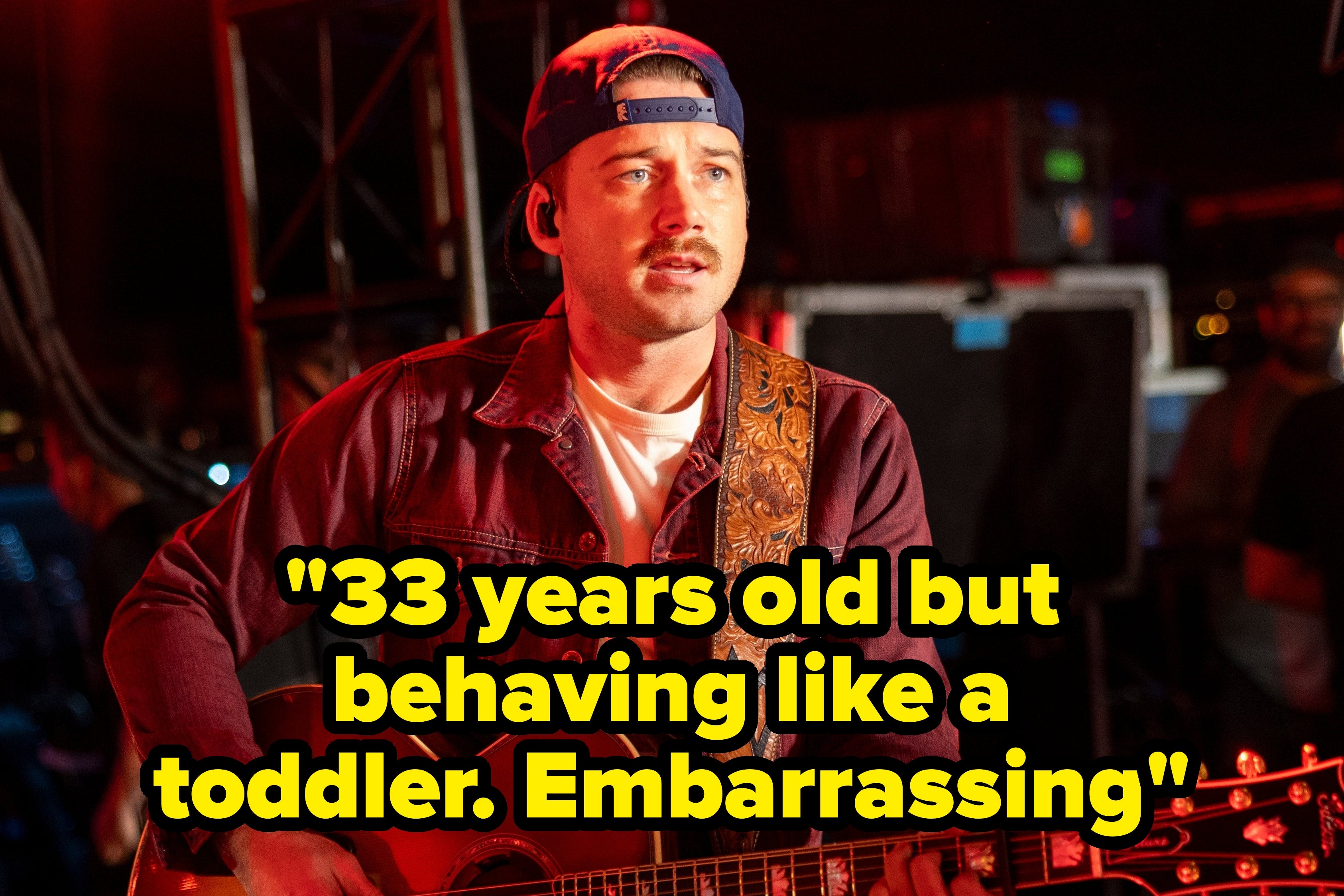 Morgan Wallen playing an acoustic guitar backstage with a casual cap and jacket, in a relaxed setting with crew members around