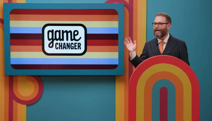Man in suit with orange tie stands next to a retro-style TV display with "Game Changer" text, smiling and waving on a vibrant set