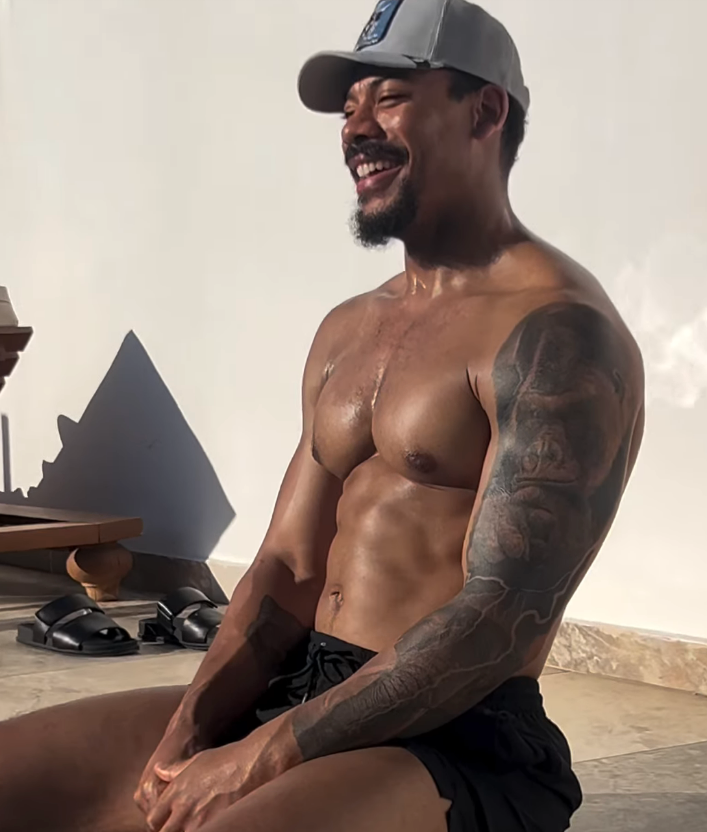 Mann som sitter uten overkropp, iført caps og shorts, smiler utendørs. Solbelyst scene