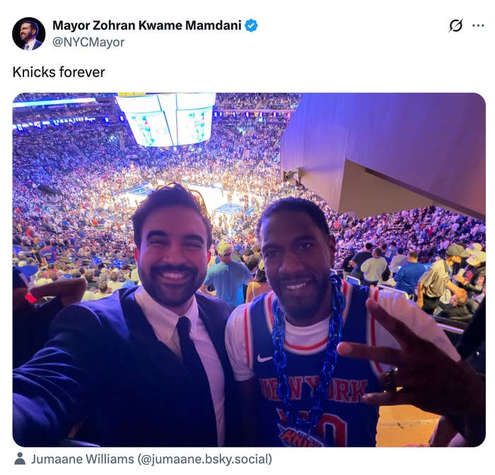 Two radical   smiling, with 1  wearing a New York Knicks jersey. They're astatine  a crowded hoops  game, taking a selfie from the stands