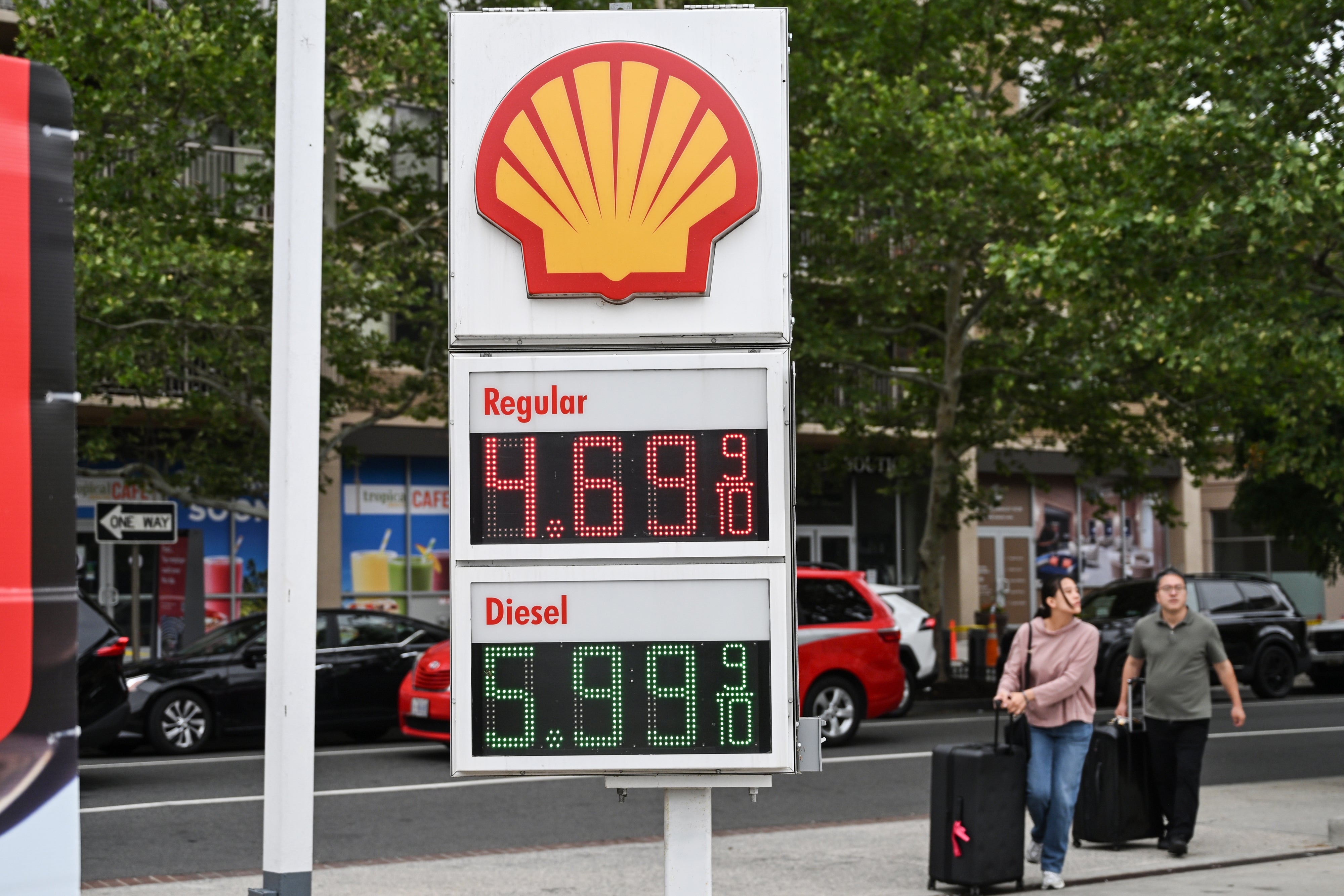 Shell bensinstasjon-skilt viser priser: Vanlig til $4,69 og Diesel til $5,99, med fotgjengere og biler i bakgrunnen