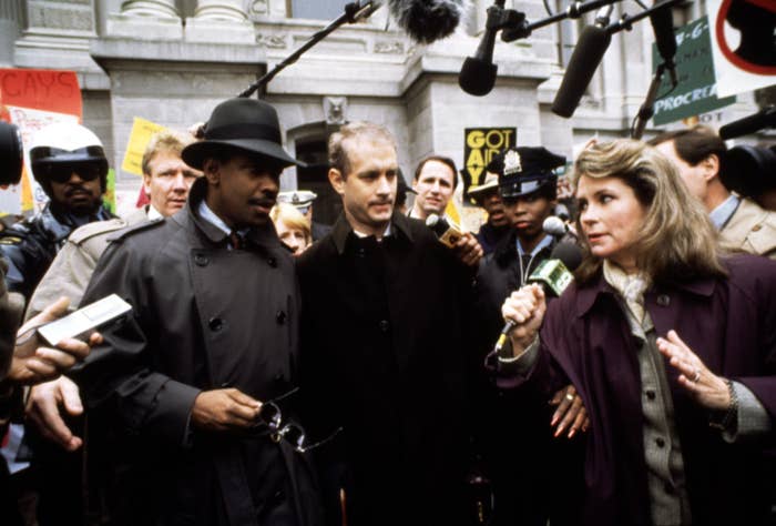 Two individuals in formal coats are surrounded by media and microphones outside a building, amid a crowd with protest signs
