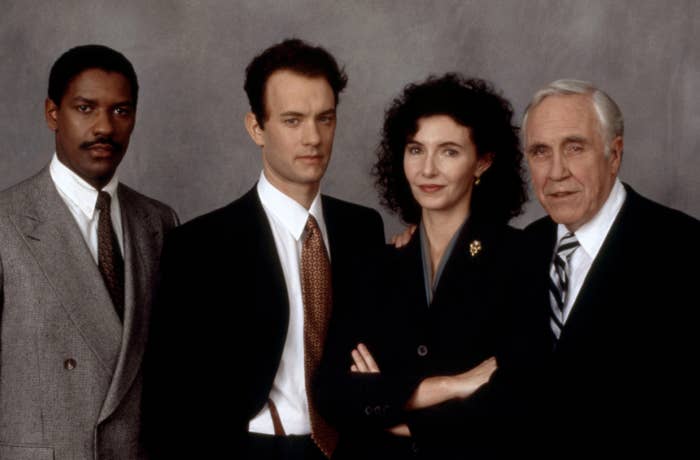 Four individuals in formal attire, possibly from a movie or TV scene, standing together against a neutral background