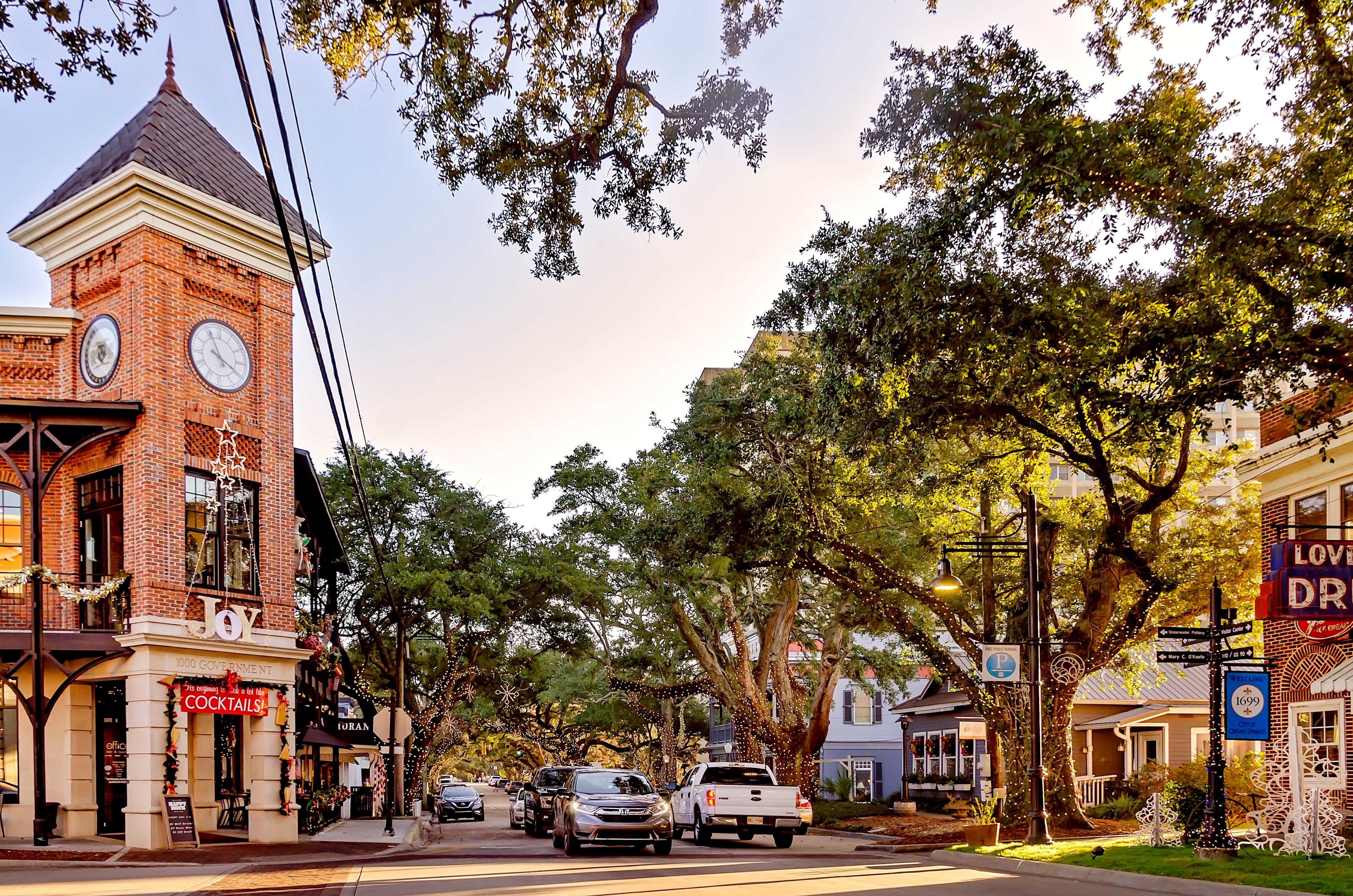 A charming thoroughfare  country   with cars parked, a ceramic  gathering  with a timepiece  tower, and ample  trees lining the street