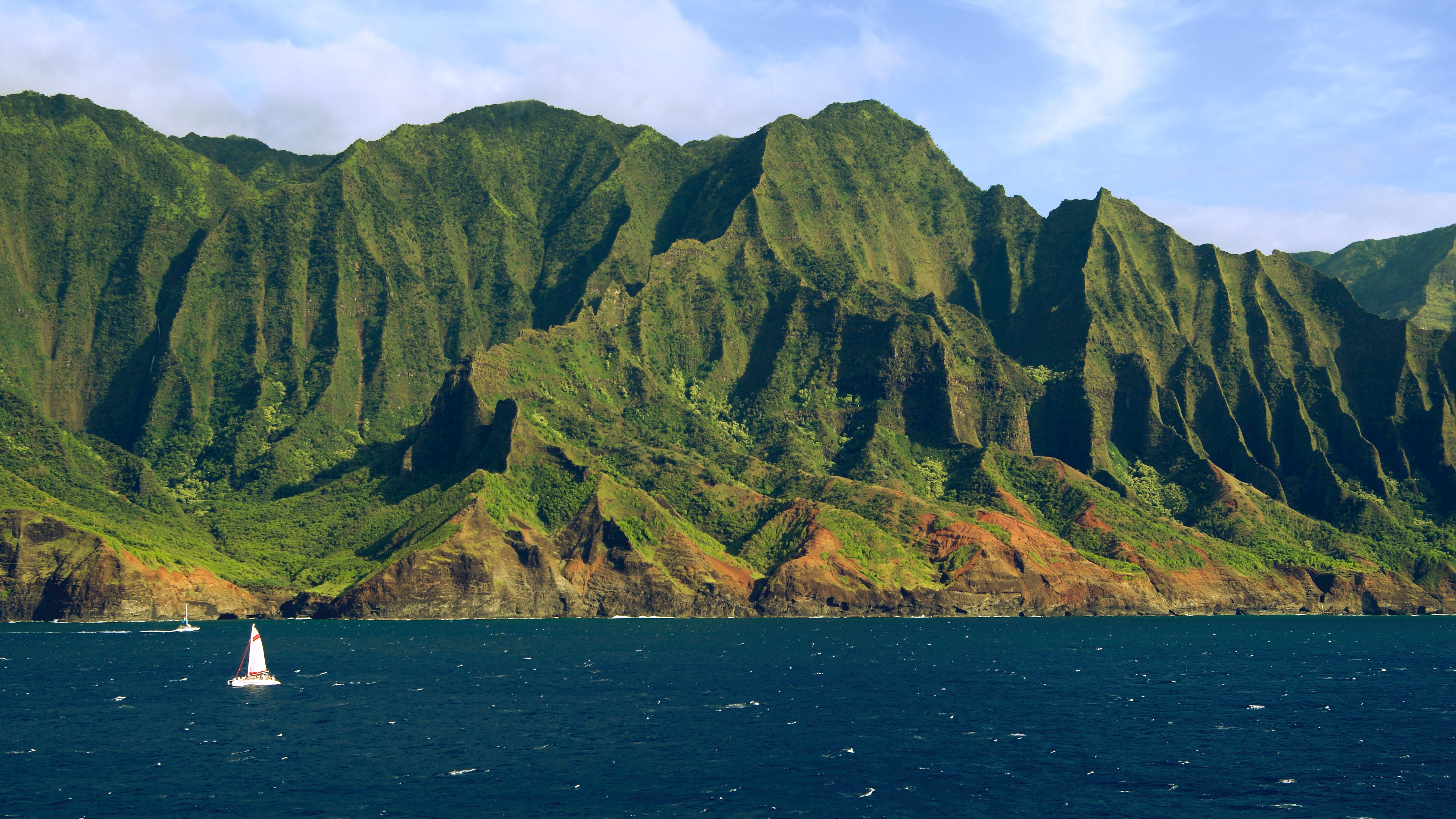 A sailboat navigates the water  adjacent   jagged, lush greenish  cliffs nether  a partially  cloudy sky