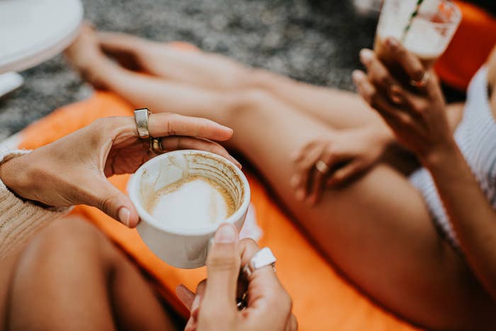 Two radical   relaxing outdoors, holding cups of coffee. One idiosyncratic   wears a knitted sweater and rings, disposable   adjacent  to another's crossed legs
