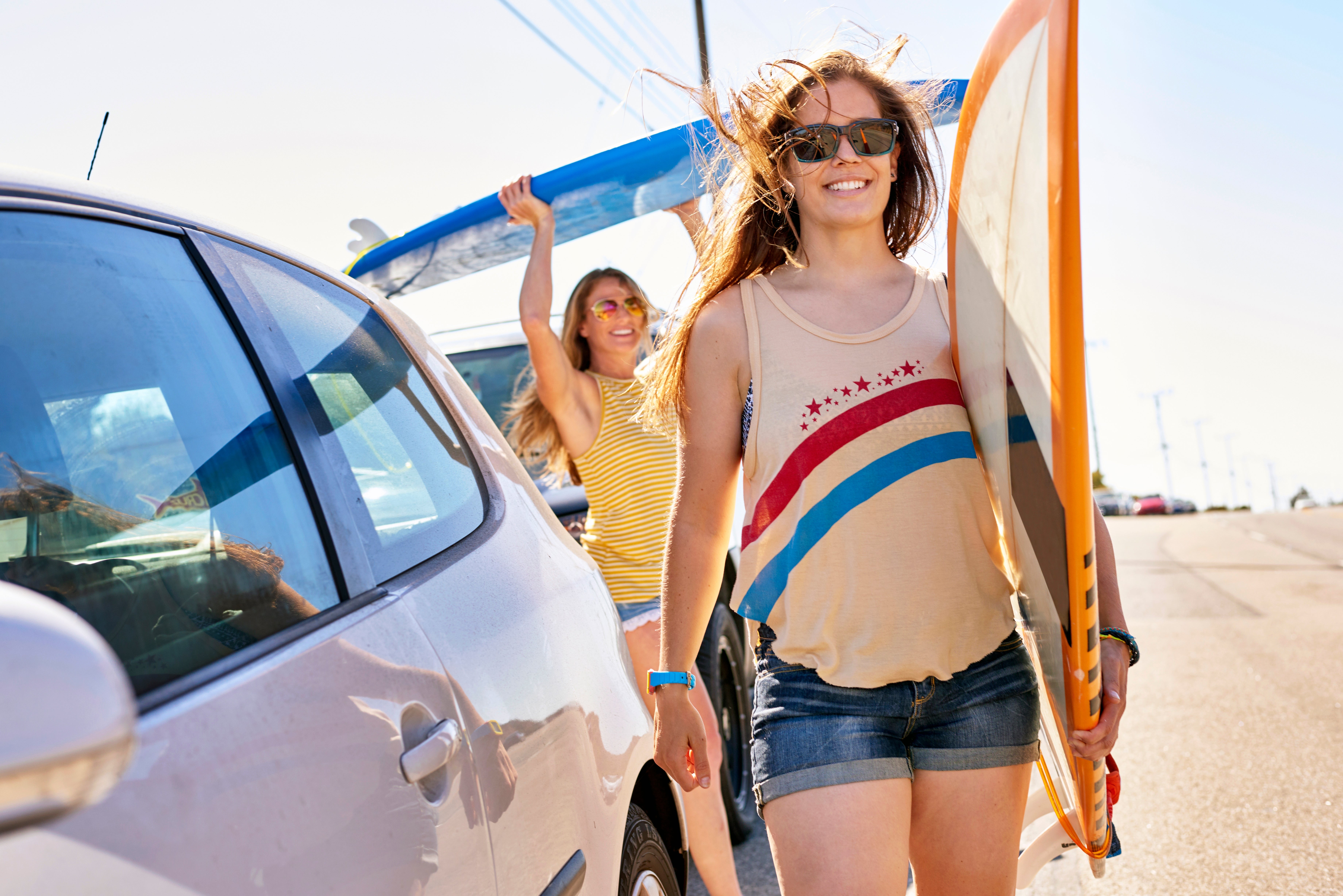 Two radical   with surfboards locomotion  beside parked cars connected  a sunny day. One idiosyncratic   wears a vessel  apical  and shorts, smiling and holding a surfboard