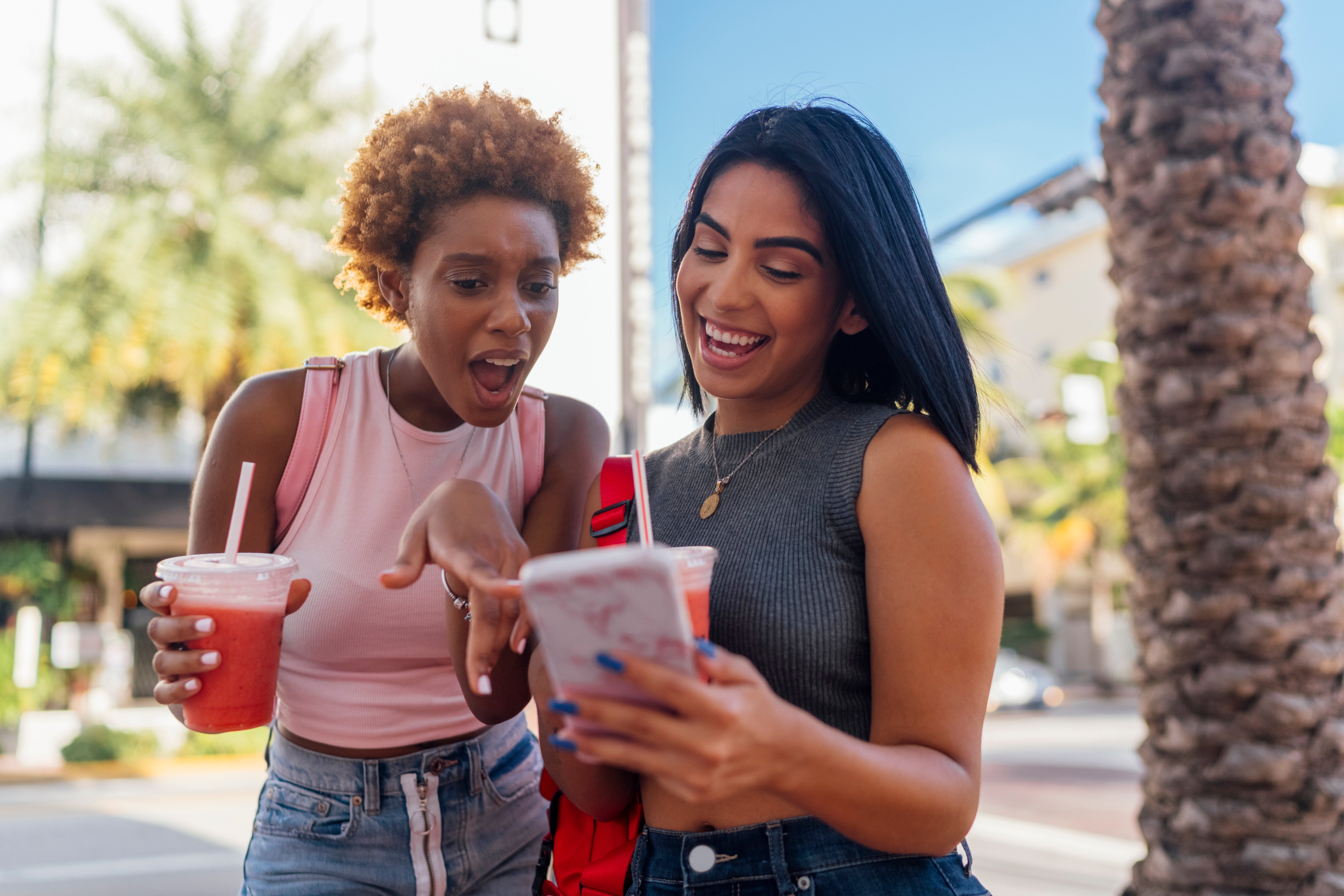Two radical   smiling and looking astatine  a phone, each   holding a drink, outdoors connected  a sunny day