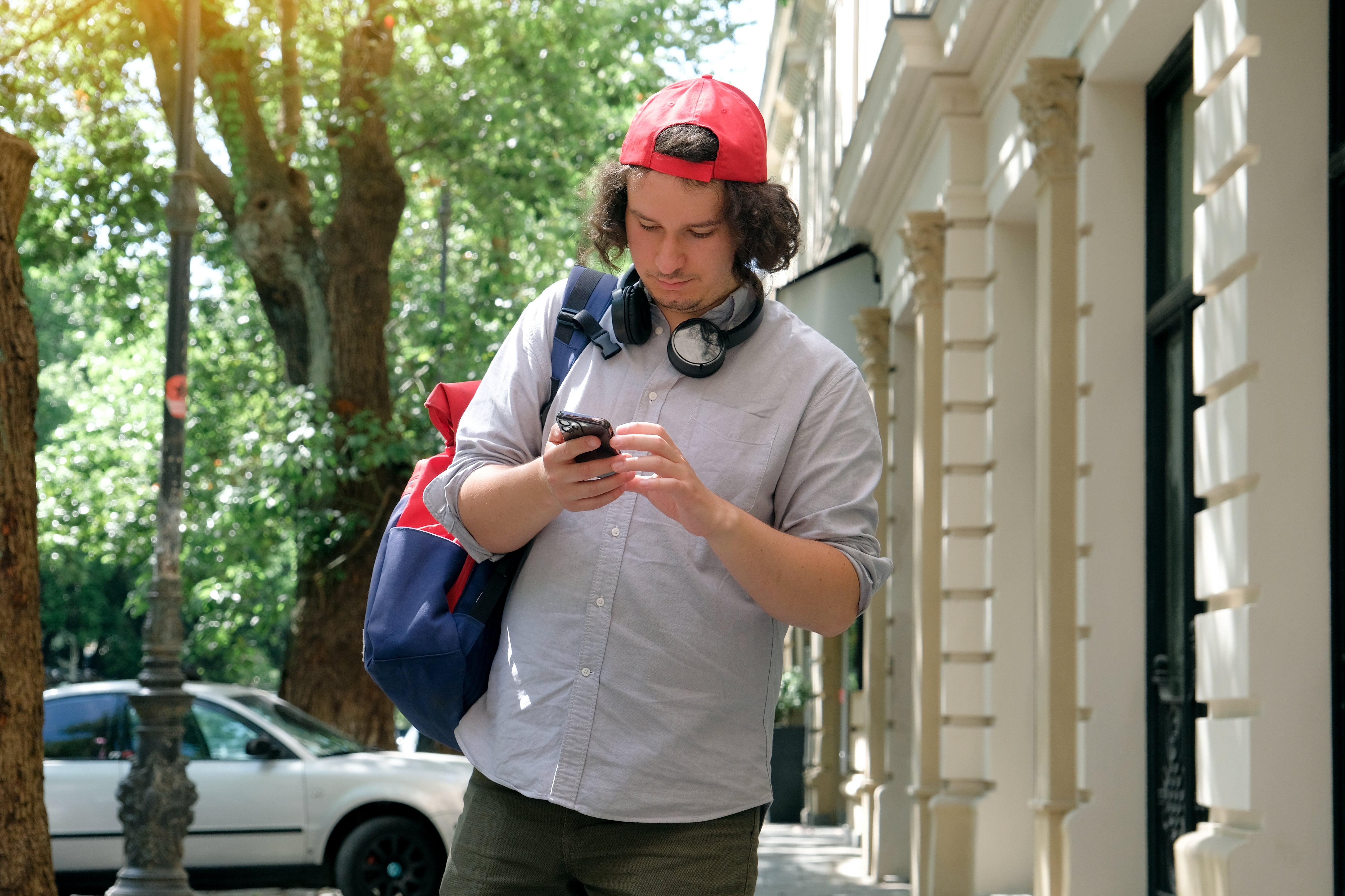 Person wearing a cap, headphones astir   neck, checks telephone  portion    walking connected  a sidewalk carrying a ample  backpack