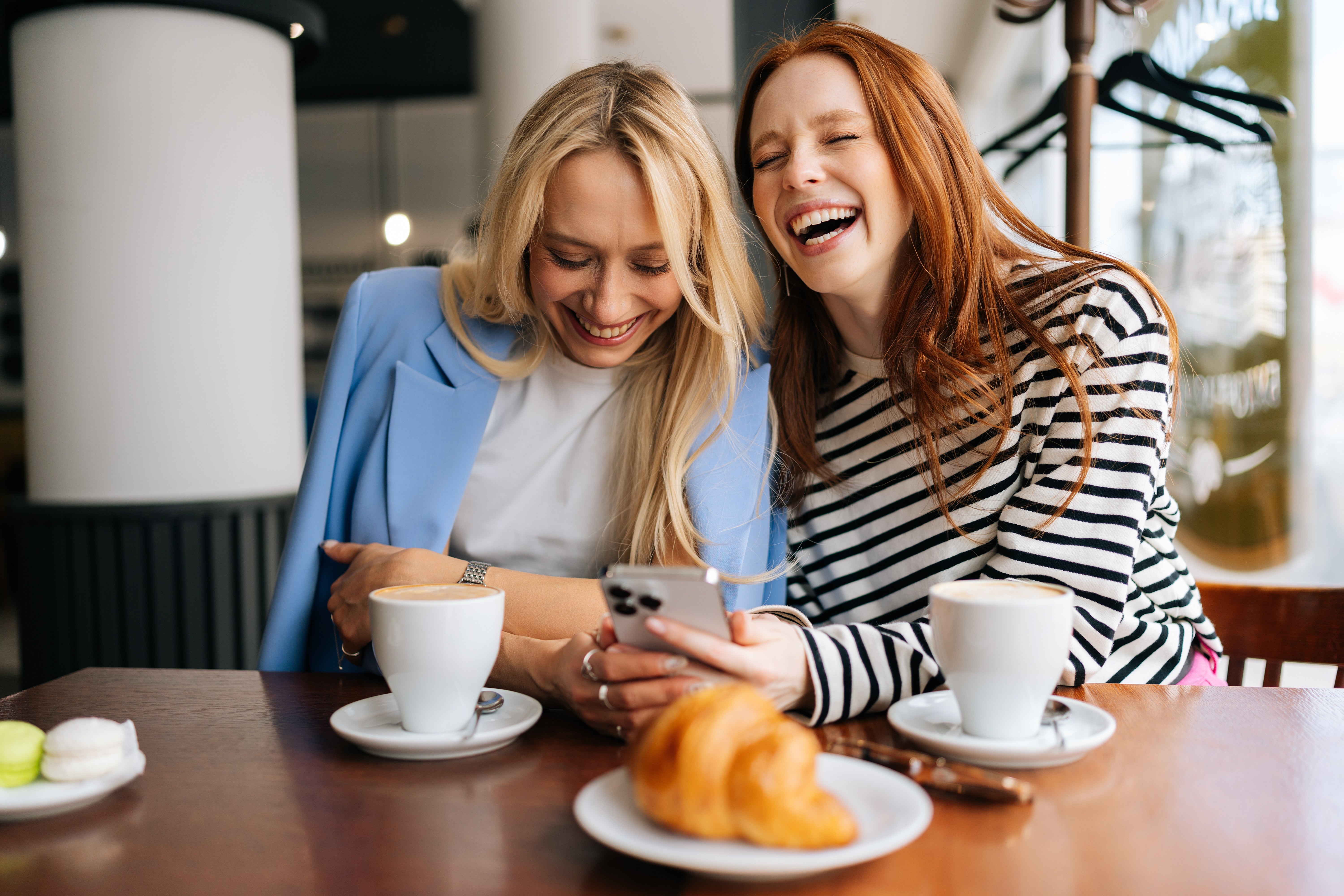 Two women laughing unneurotic  astatine  a café table, looking astatine  a phone, with java  and a croissant successful  beforehand   of them