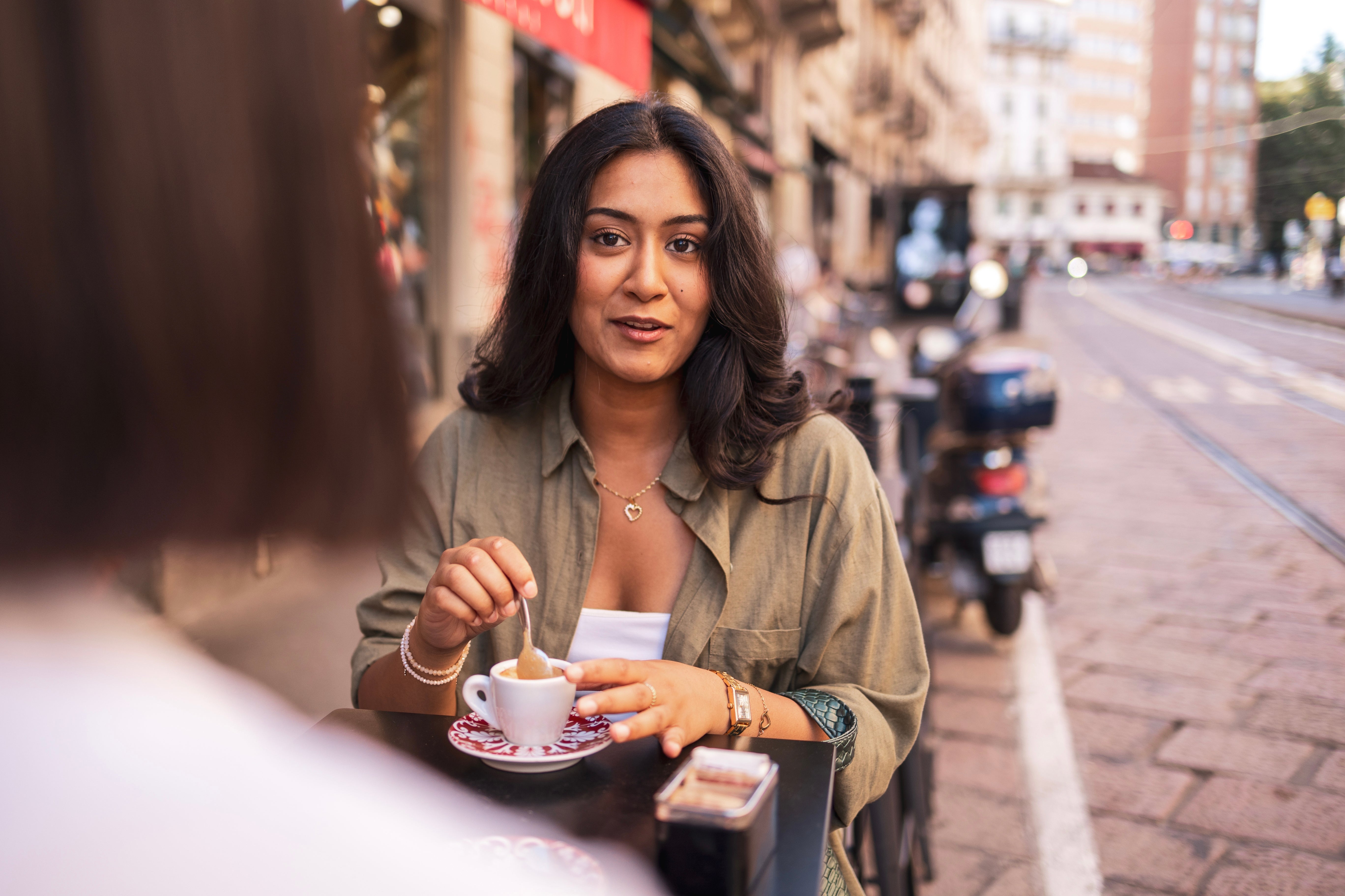 Person sitting astatine  an outdoor cafe table, holding a java  cupful  and smiling, with a metropolis  thoroughfare  successful  the background