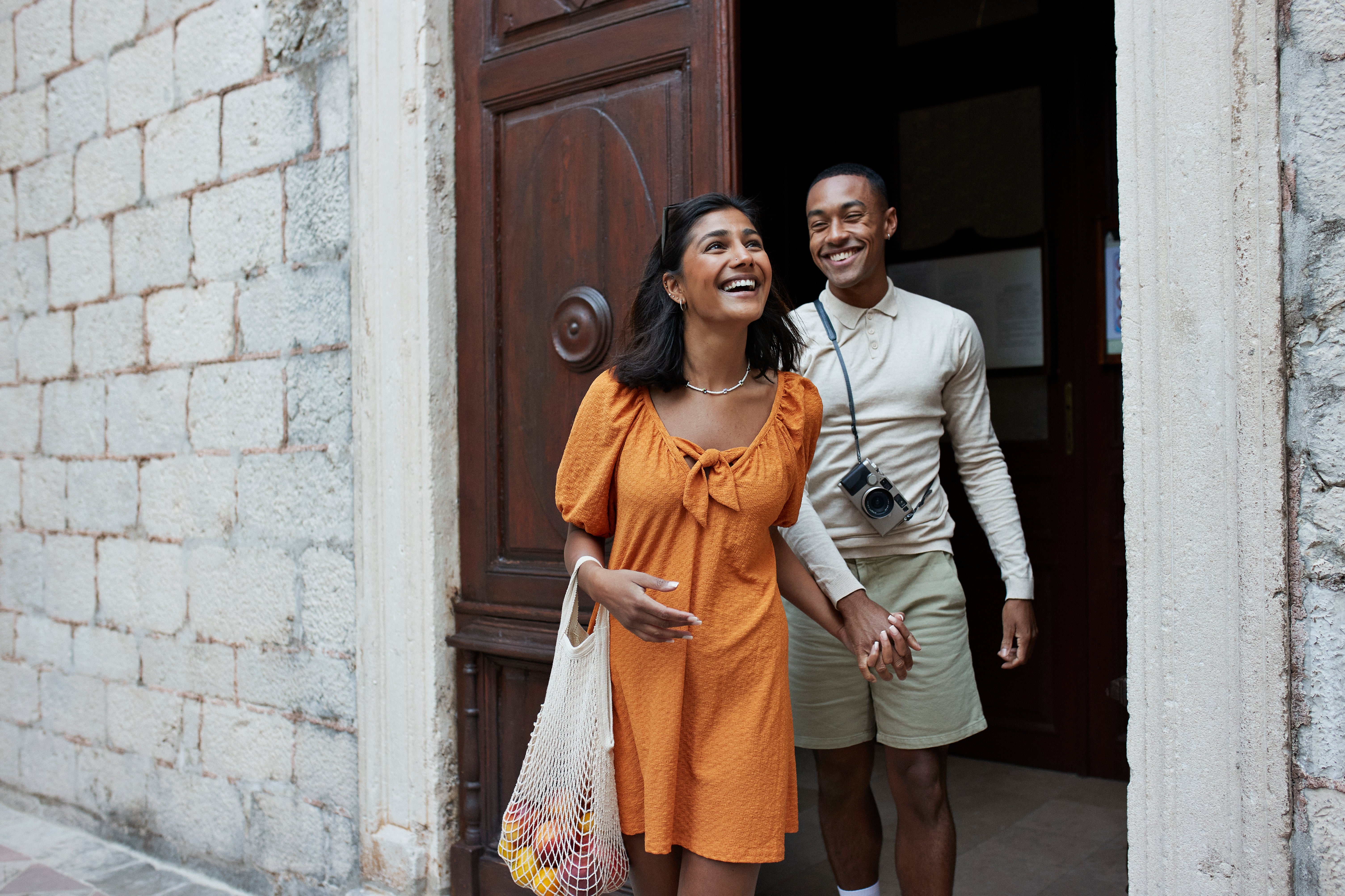 Two radical   happily exit a building, holding hands. One is wearing a knee-length formal  and the different   wears a long-sleeve garment  with shorts and a camera