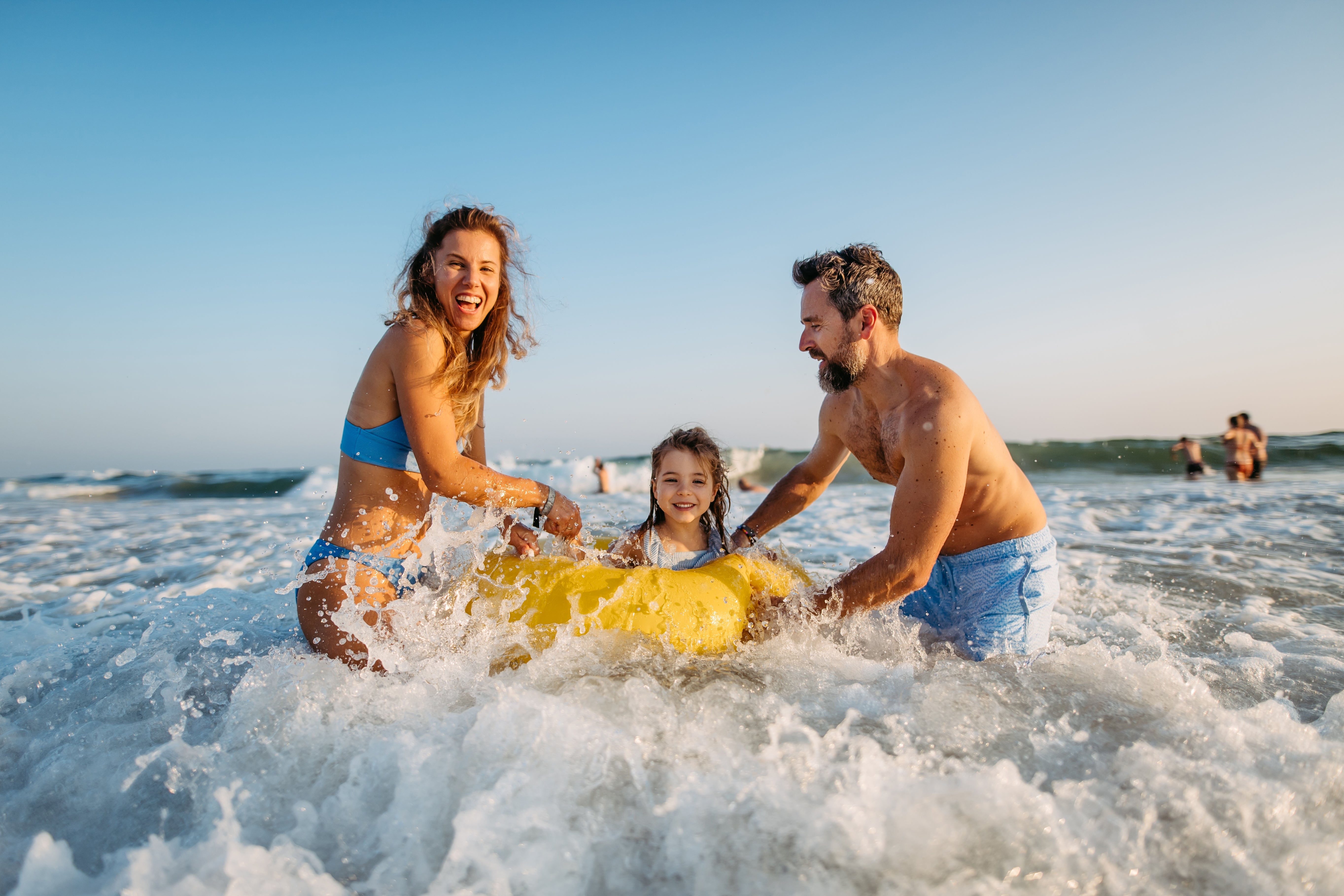A household  of 3  plays joyfully successful  water  waves, holding onto a yellowish  inflatable raft, with smiles and bedewed  hair