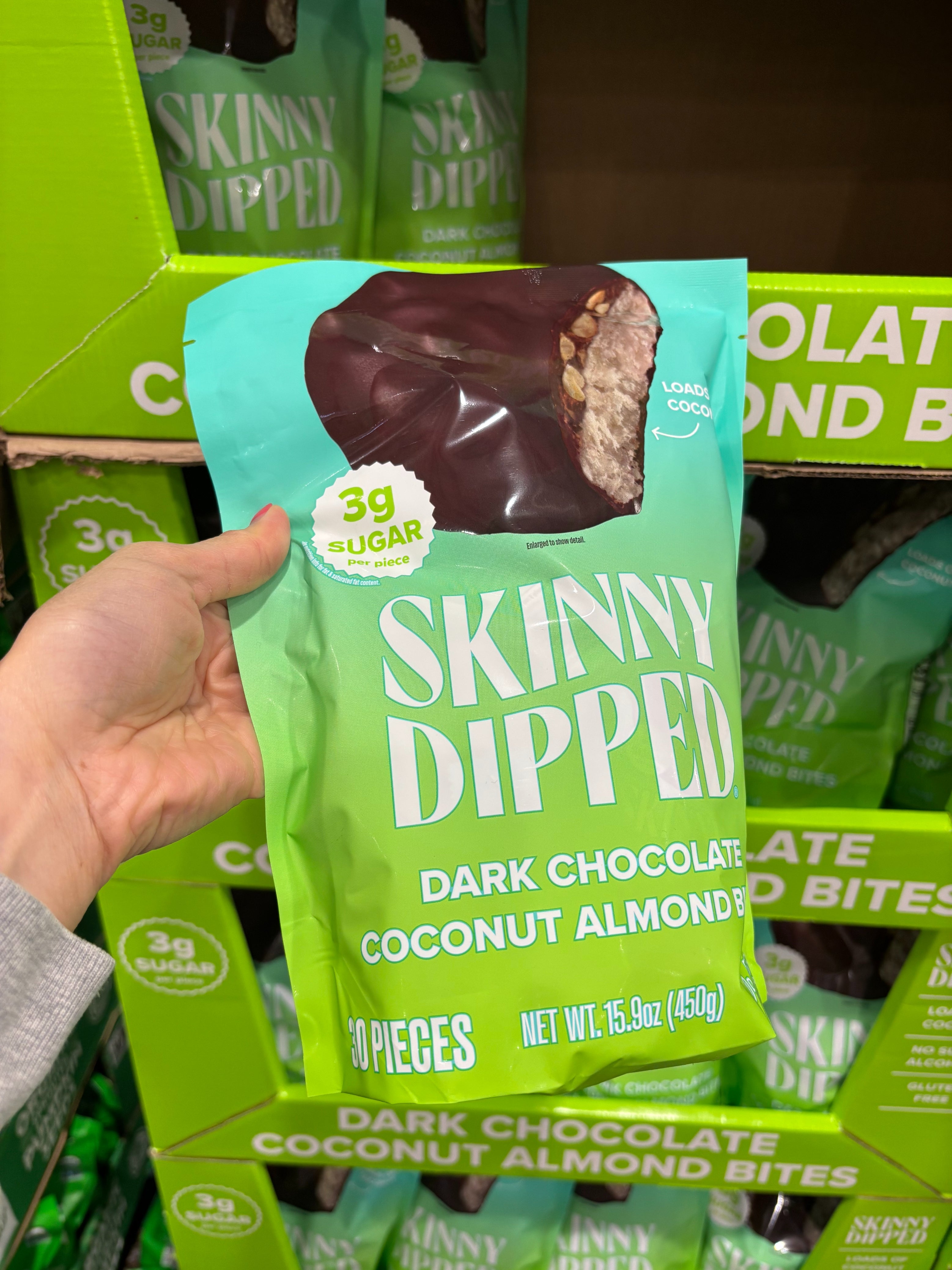 A hand holds a bag of Skinny Dipped dark chocolate coconut almond bites with packaging emphasizing 3g sugar per piece