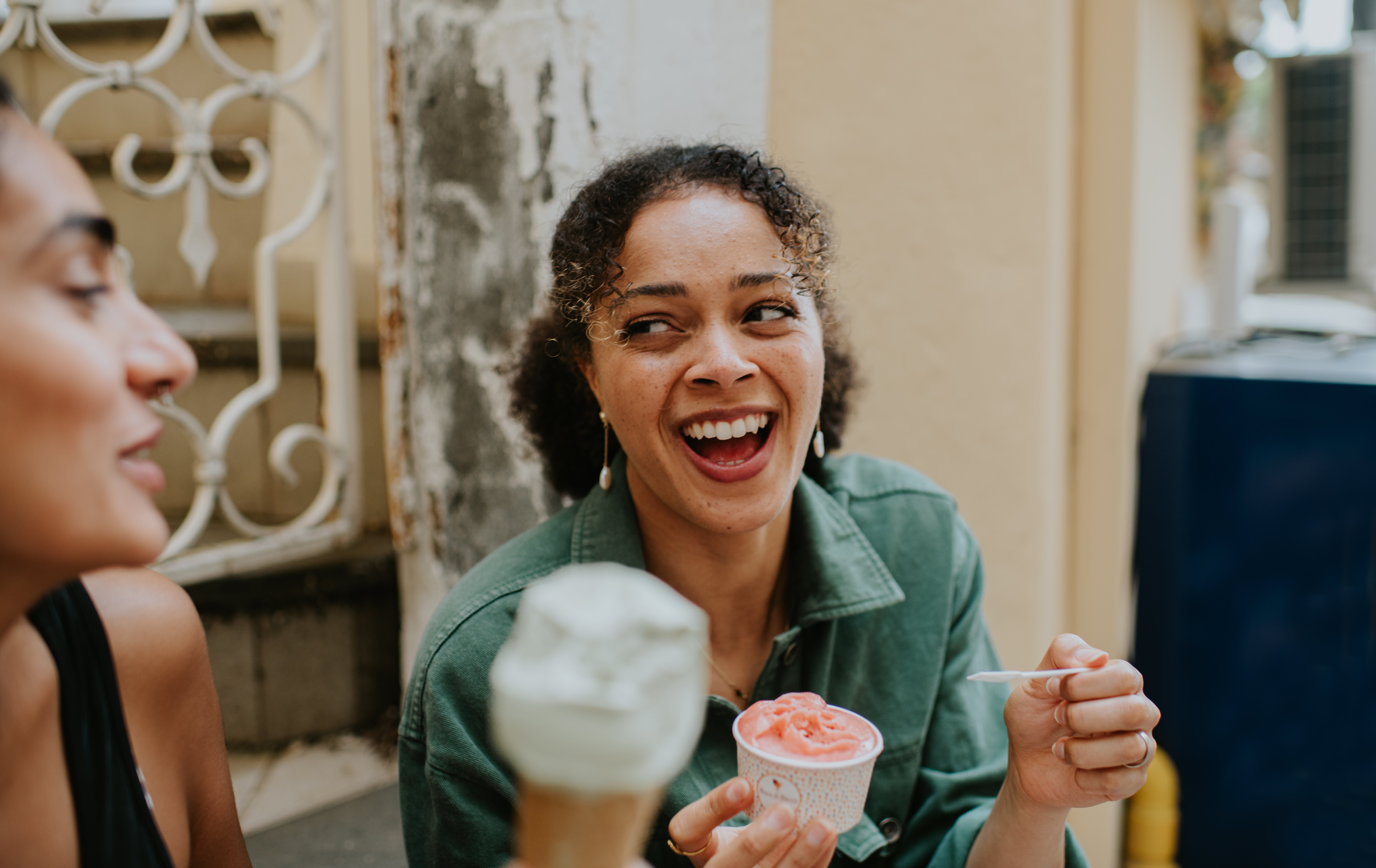 Woman laughing and holding a cupful  of gelato, sitting extracurricular  with a friend