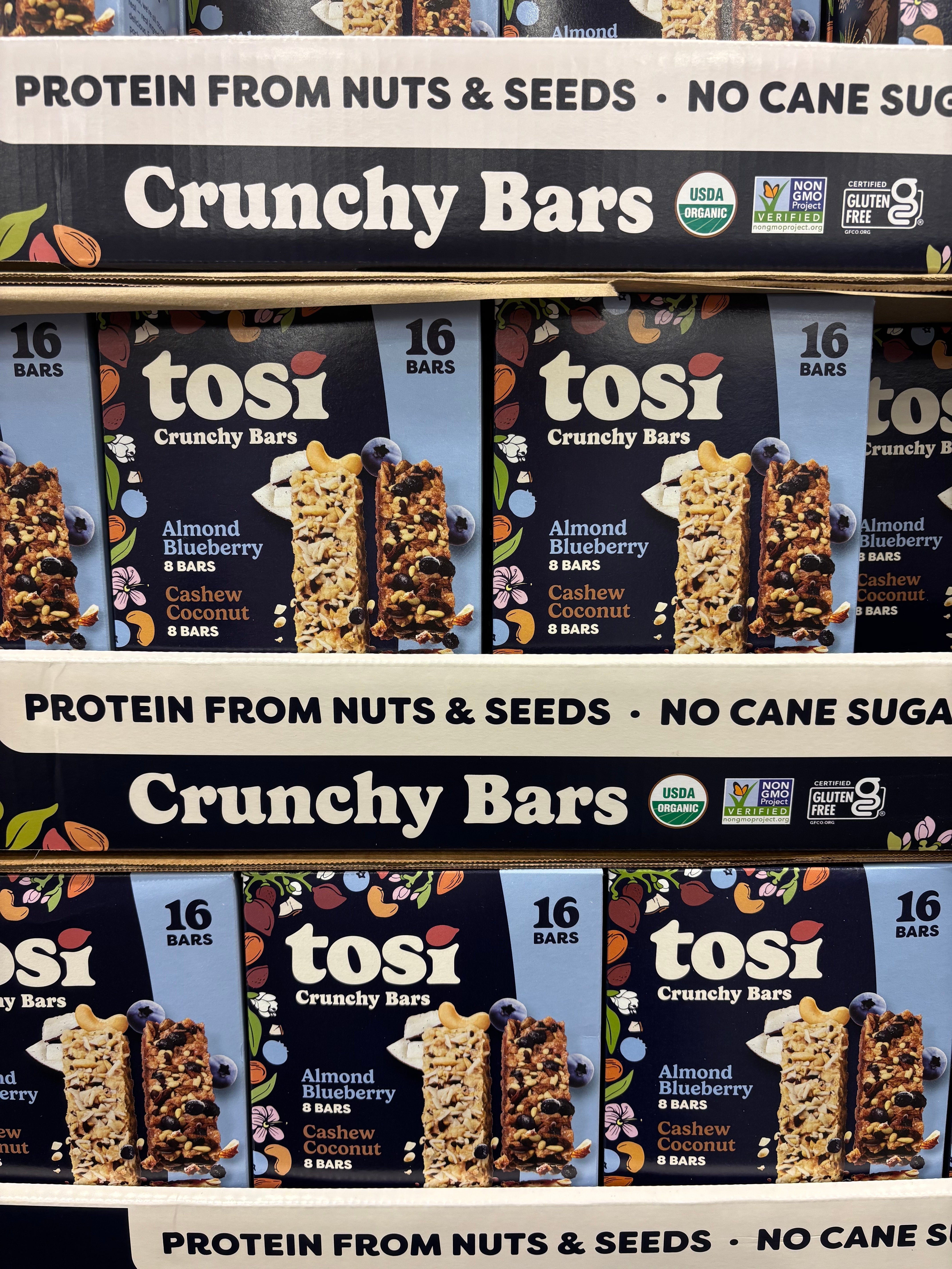 Tosi Crunchy Bars packaging display with almond blueberry and cashew flavors, highlighting "protein from nuts &amp;amp; seeds, no cane sugar."
