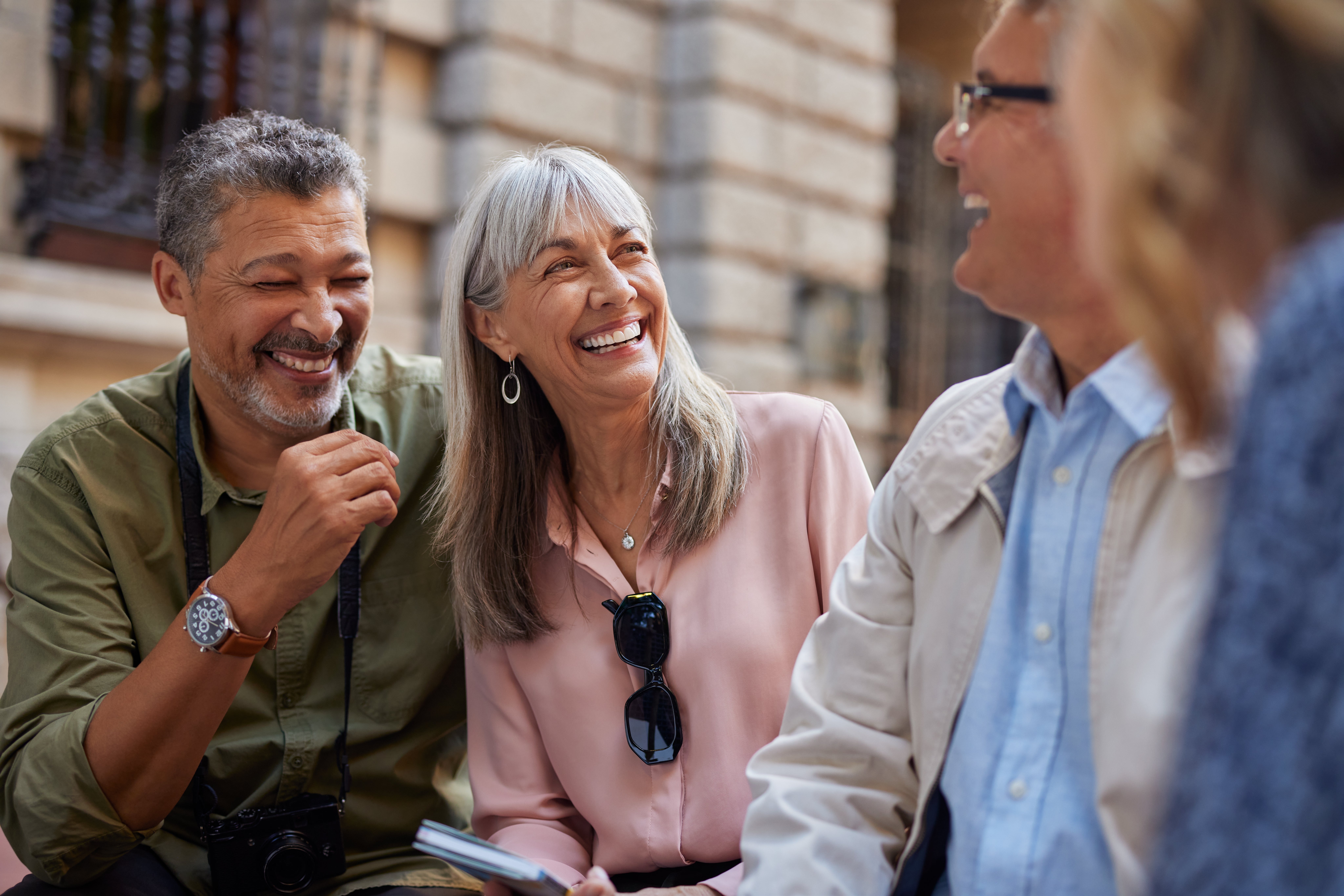 Group of 4  older adults smiling and chatting outdoors, 1  with a camera astir   their neck