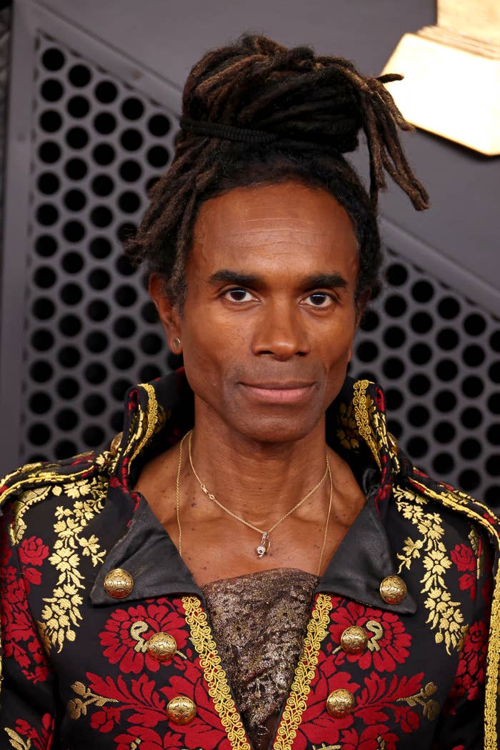 Person in elaborate floral-patterned coat with gold detailing and high collar, wearing layered necklaces. Hair styled in locs with an updo