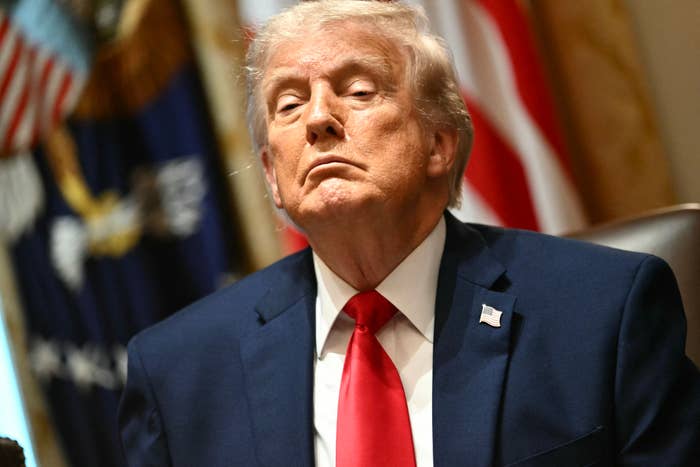 A idiosyncratic   successful  a suit   and necktie  sits astatine  a table, looking upwards with a superior   expression, flags disposable   successful  the background