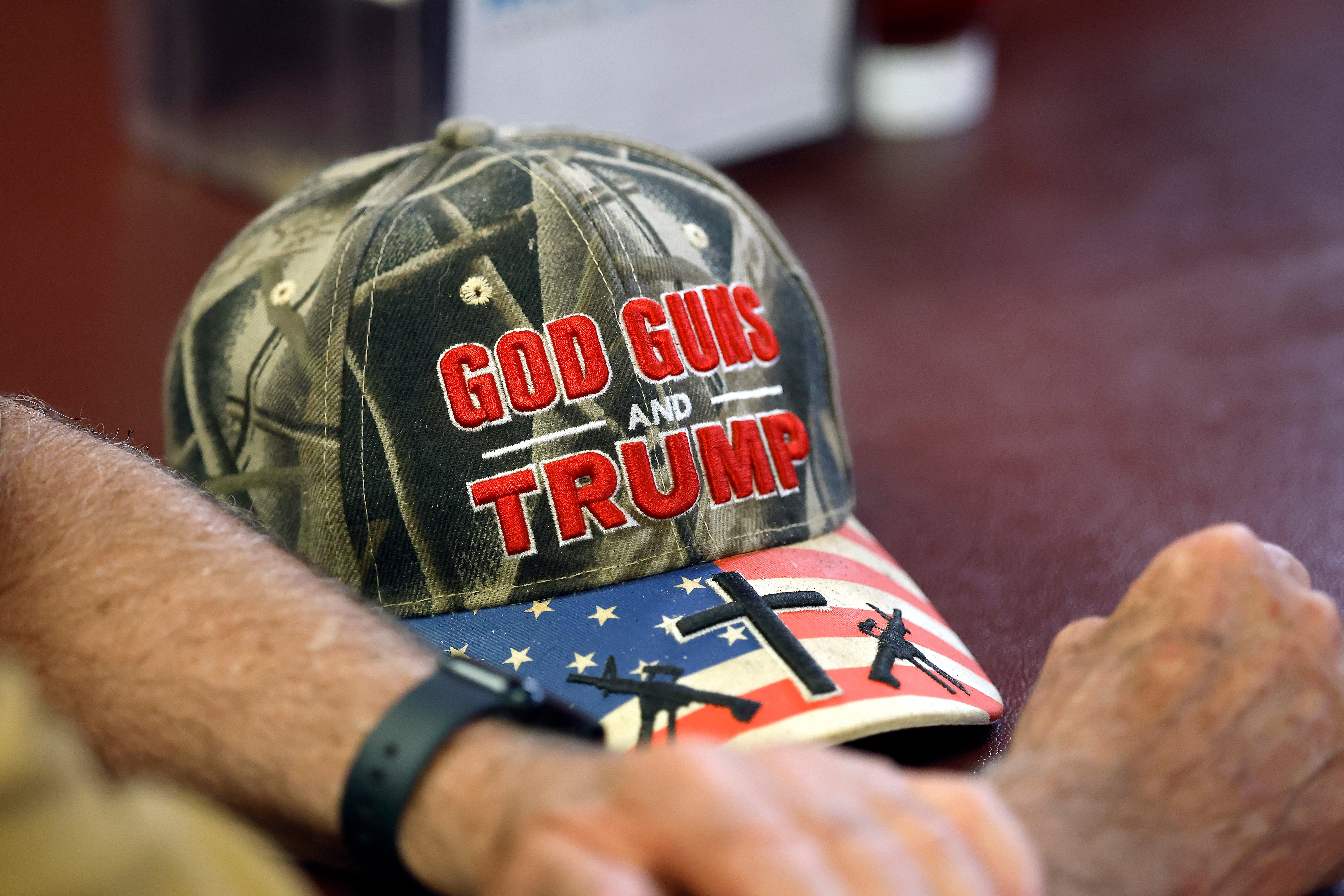 Cap with "God, Guns, and Trump" substance lies connected a array adjacent to a person's arm. The headdress features a signifier with firearms and the American flag
