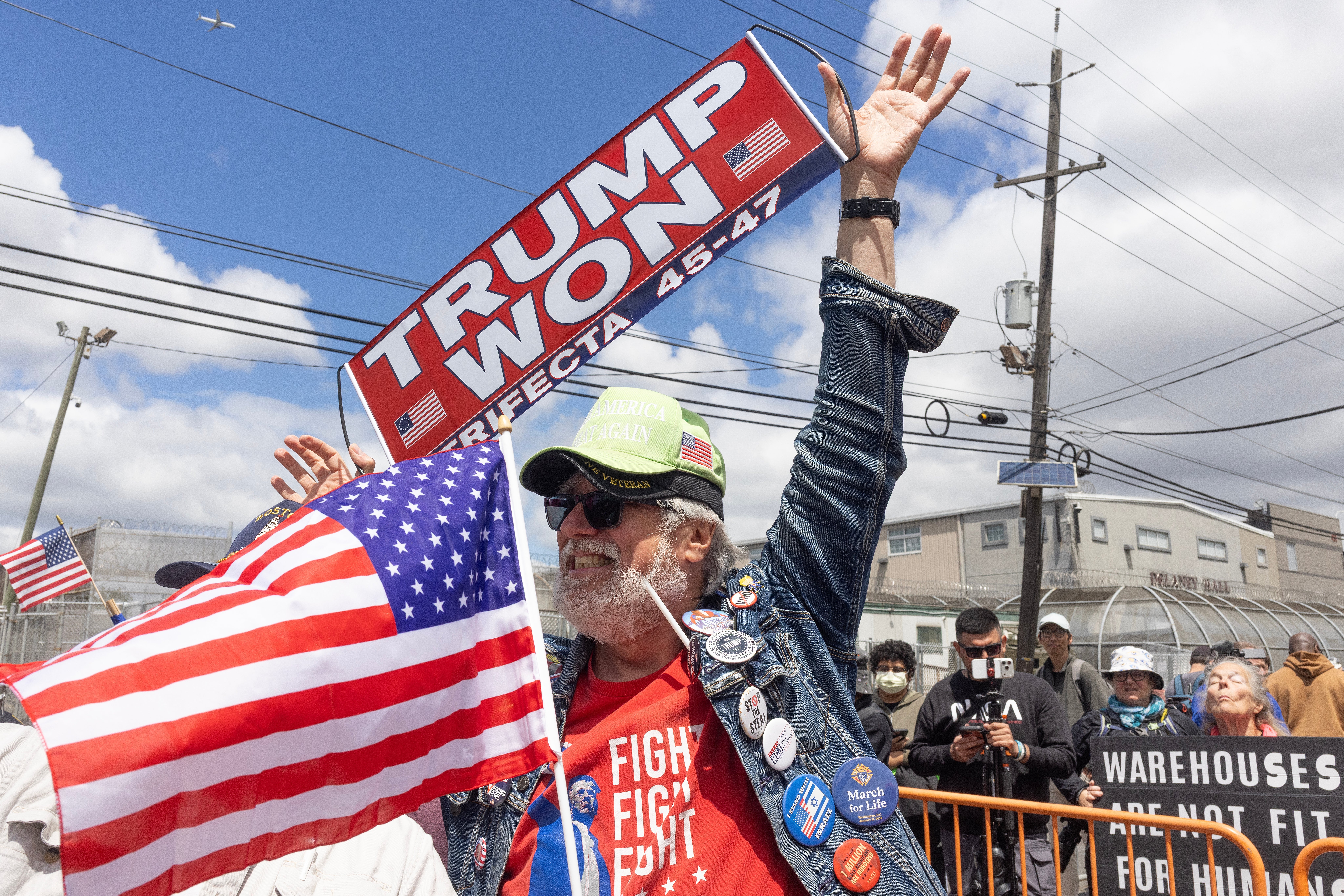 Man astatine a rally holding a "Trump Won" motion wears a denim overgarment with badges and a cap, waving an American flag. Others stitchery down a fence