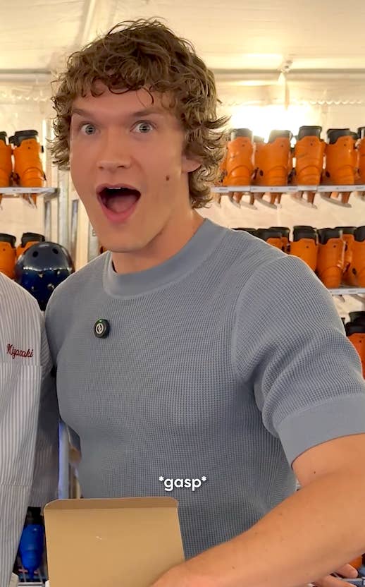 Person with curly hair, wearing a short-sleeve knit top, looks surprised with a wide-eyed expression in front of a wall of roller skates