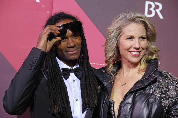 Two individuals smiling on a red carpet. The person on the left is adjusting sunglasses while wearing a tuxedo. The person on the right is in a bejeweled jacket