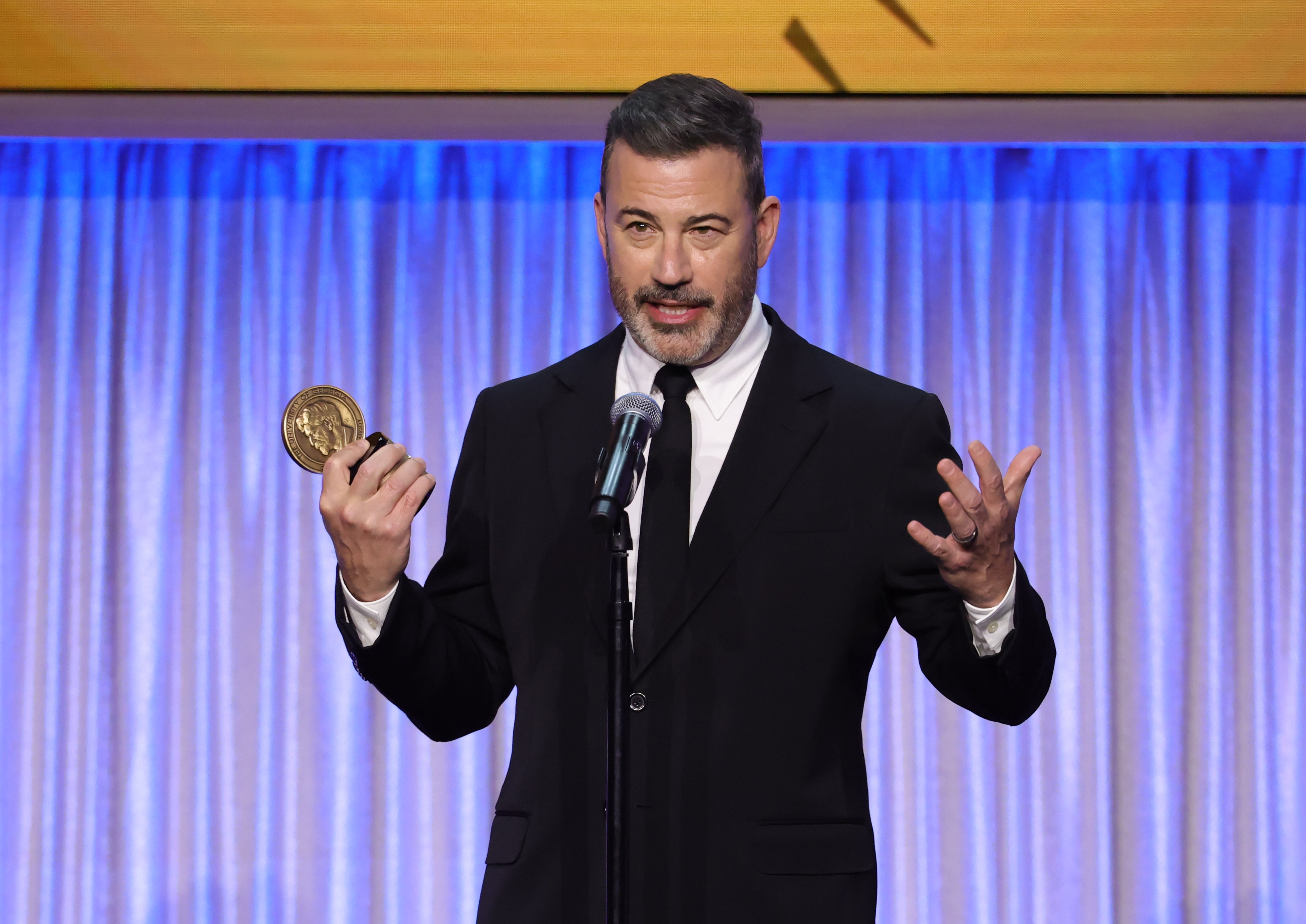 A antheral   successful  a suit   holding an award, speaking into a microphone connected  signifier    with curtains down  him