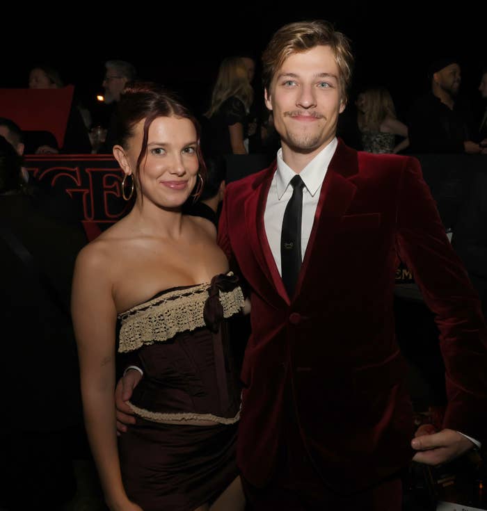 Two people pose on the red carpet. The woman wears a strapless dress with lace trim; the man wears a velvet suit with a tie