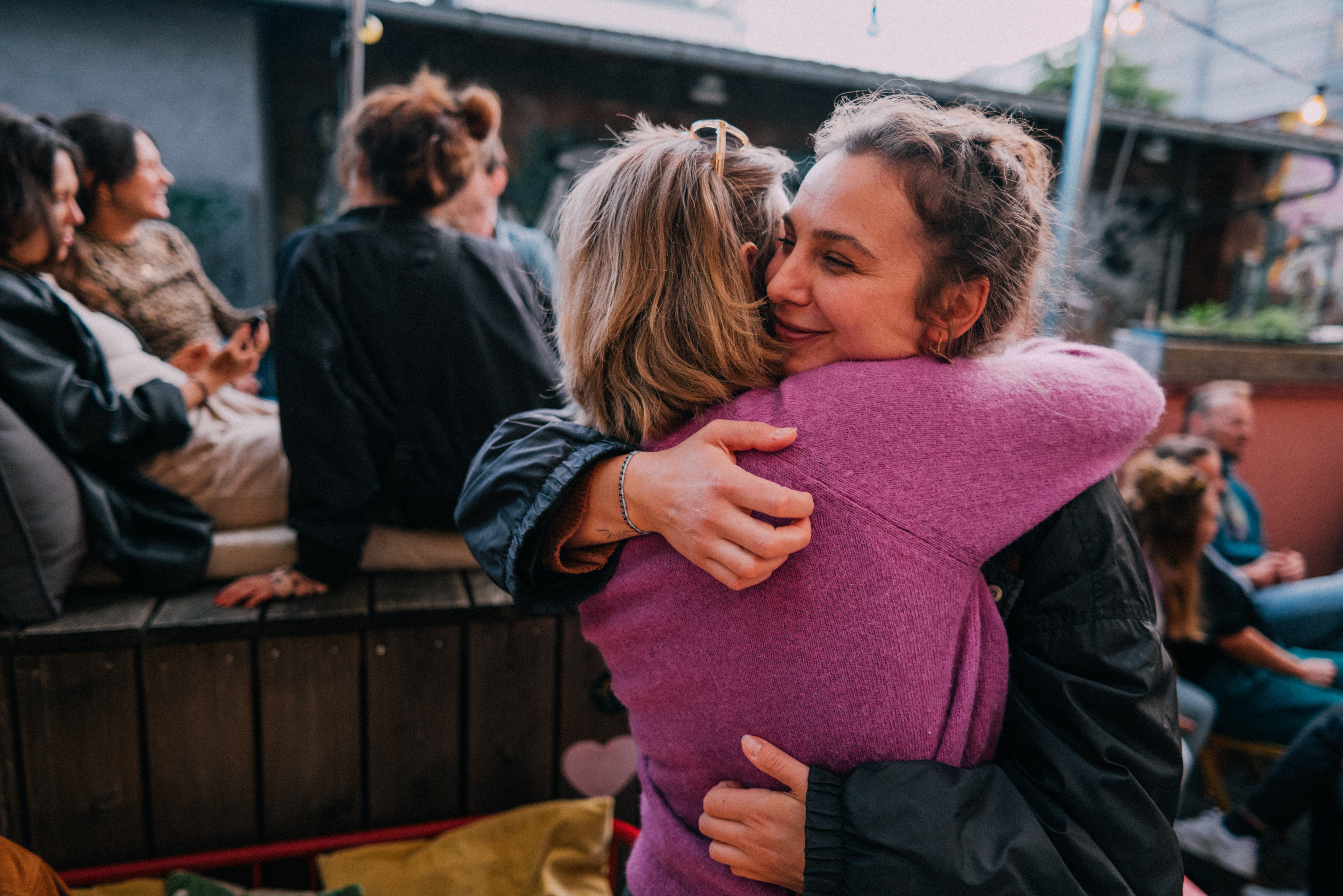 Two radical hugging warmly successful a casual outdoor setting, others sitting and chatting successful the background