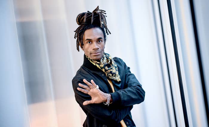 A person in a stylish jacket and patterned scarf, with dreadlocks tied up, gesturing expressively in a modern setting