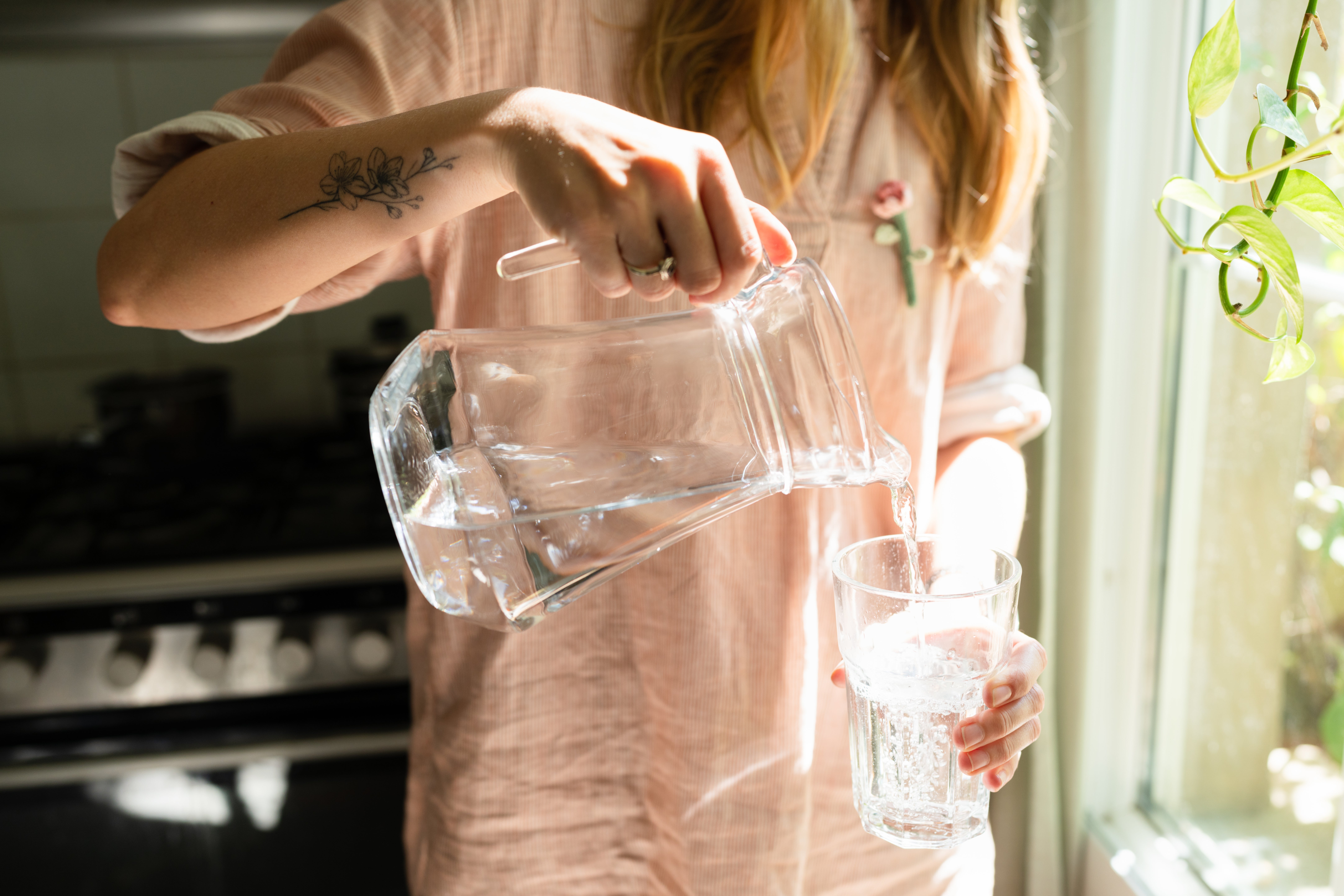 Person successful  casual covering  pouring h2o  from a solid  pitcher into a solid  by a sunlit window, with a disposable   floral tattoo connected  their forearm