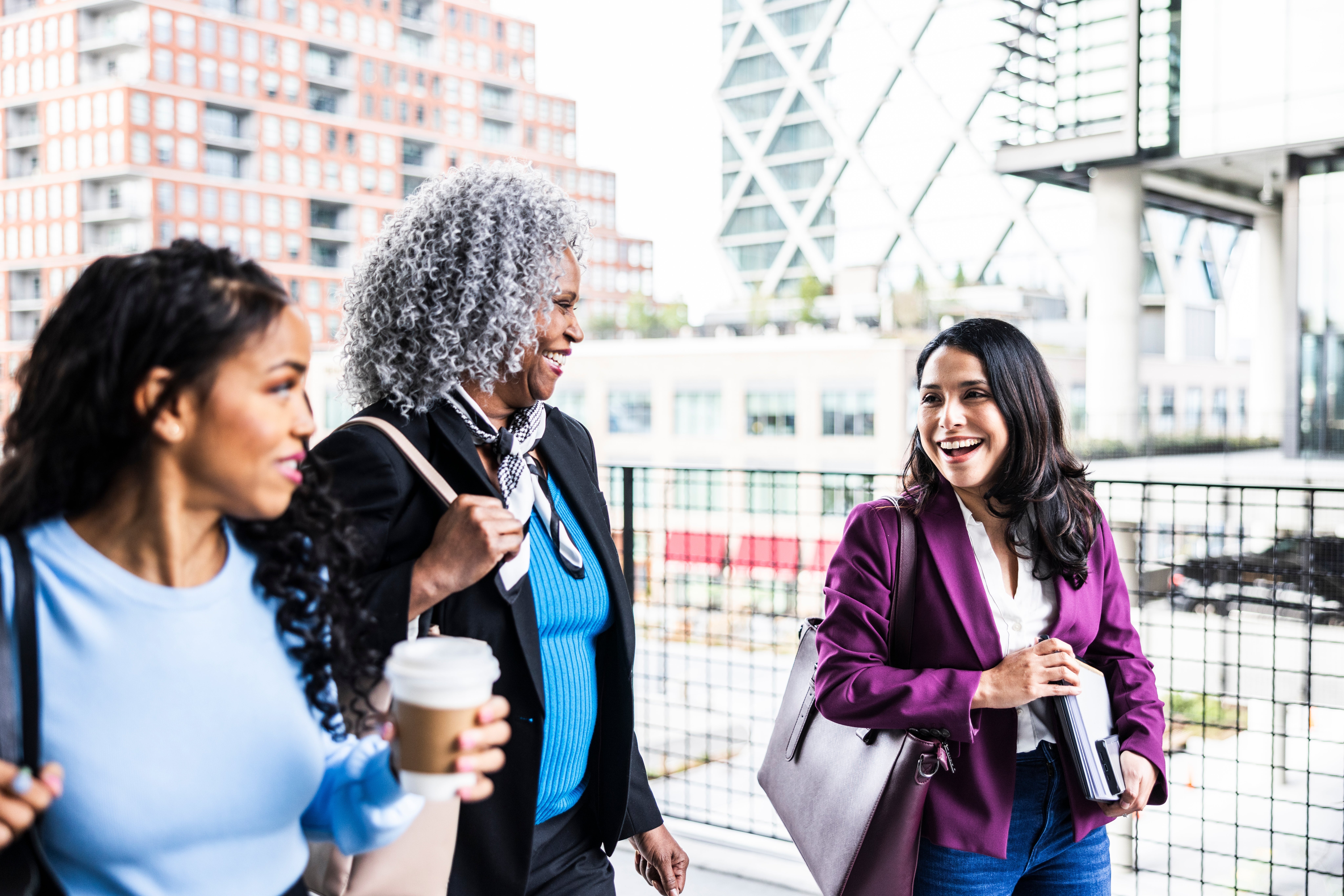 Three women locomotion  outdoors, smiling and holding java  and bags, engaging successful  conversation. Urban inheritance  with buildings and a span  visible