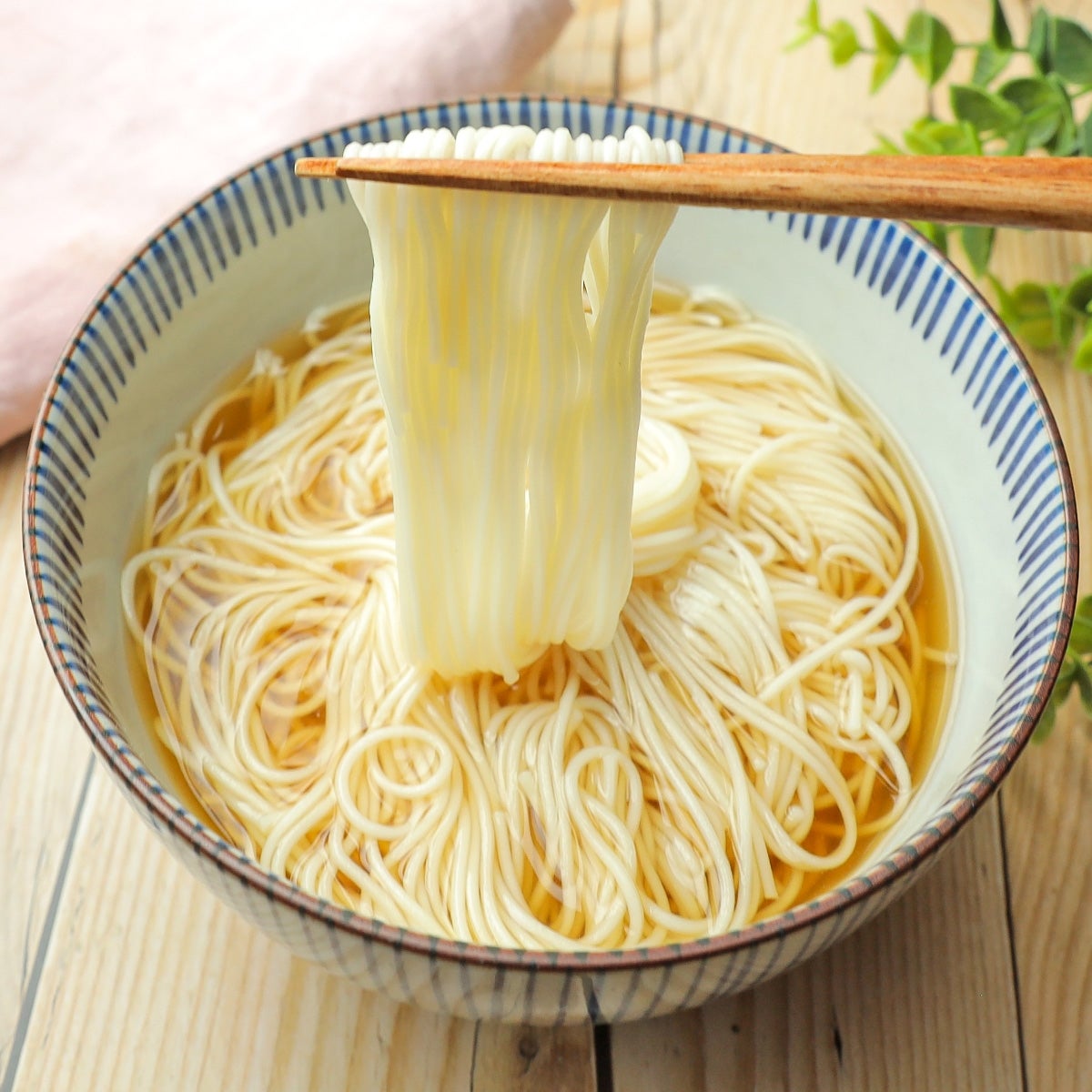 箸で持ち上げられたそうめんが器にたっぷりと盛られている。木製のテーブルの上に置かれている。