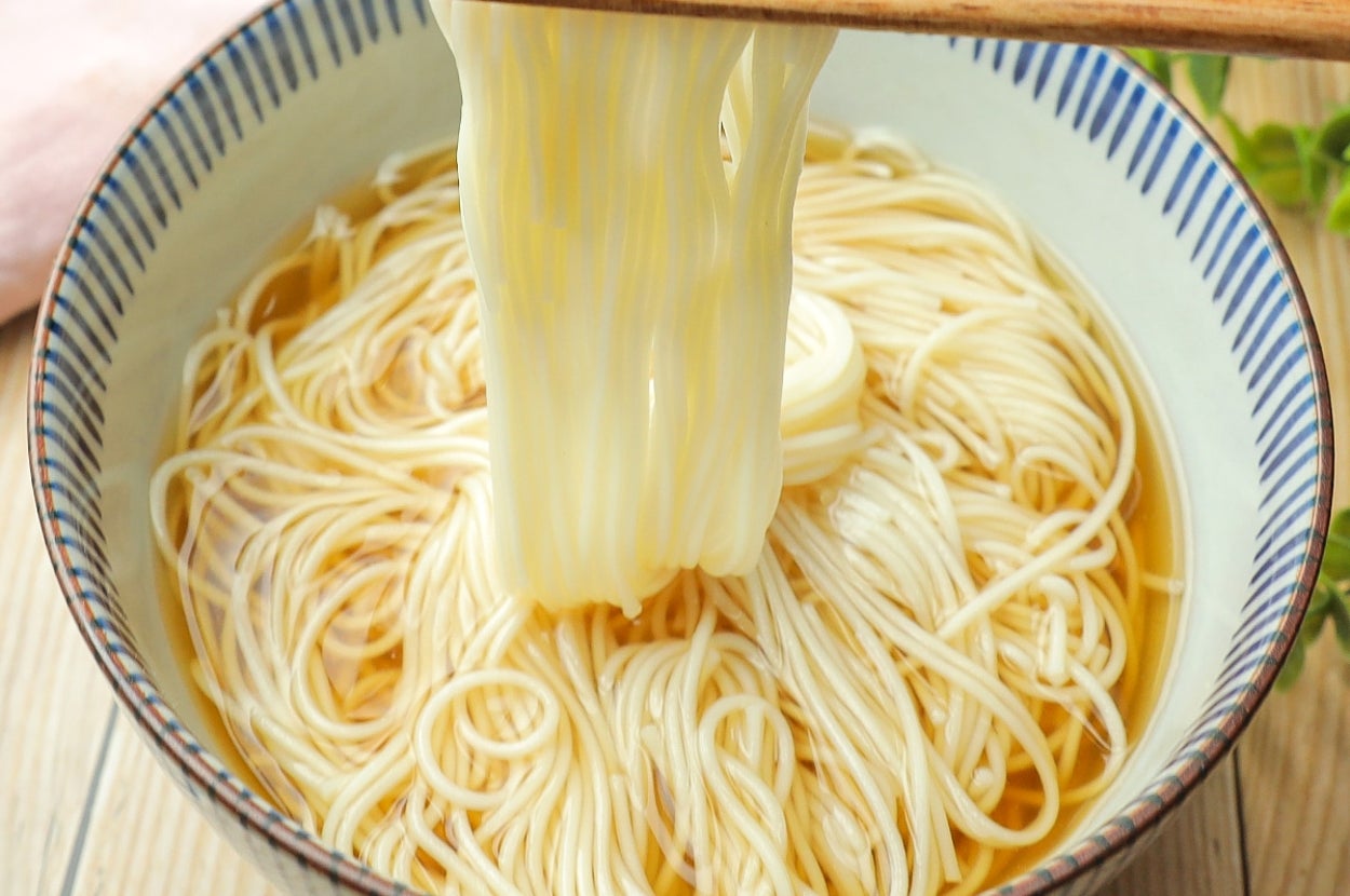 箸で持ち上げられたそうめんが器にたっぷりと盛られている。木製のテーブルの上に置かれている。
