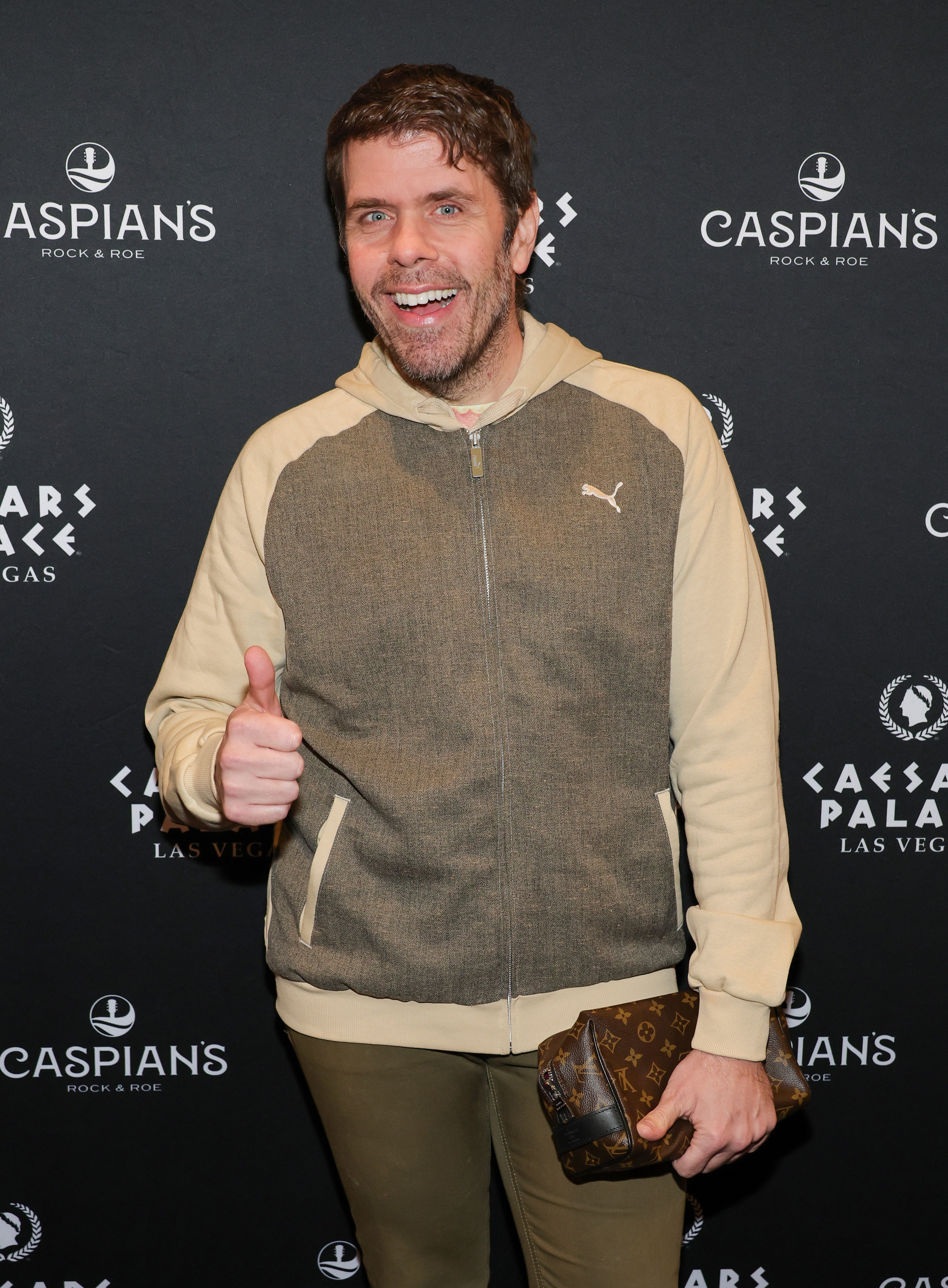 Person smiling, giving a thumbs-up connected the reddish carpet, wearing a casual hoodie and pants, holding a tiny bag