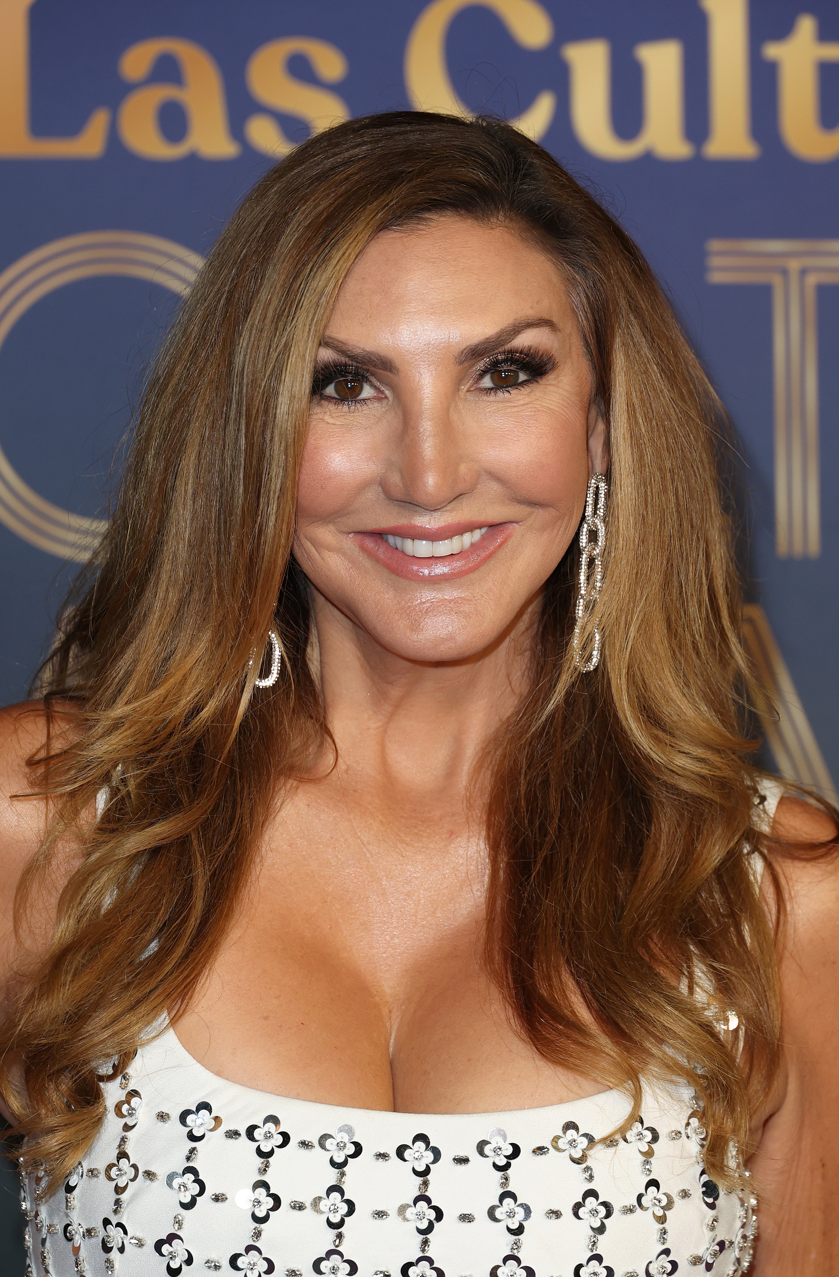 Person connected reddish carpet successful a sleeveless, patterned formal with agelong earrings, smiling astatine the camera