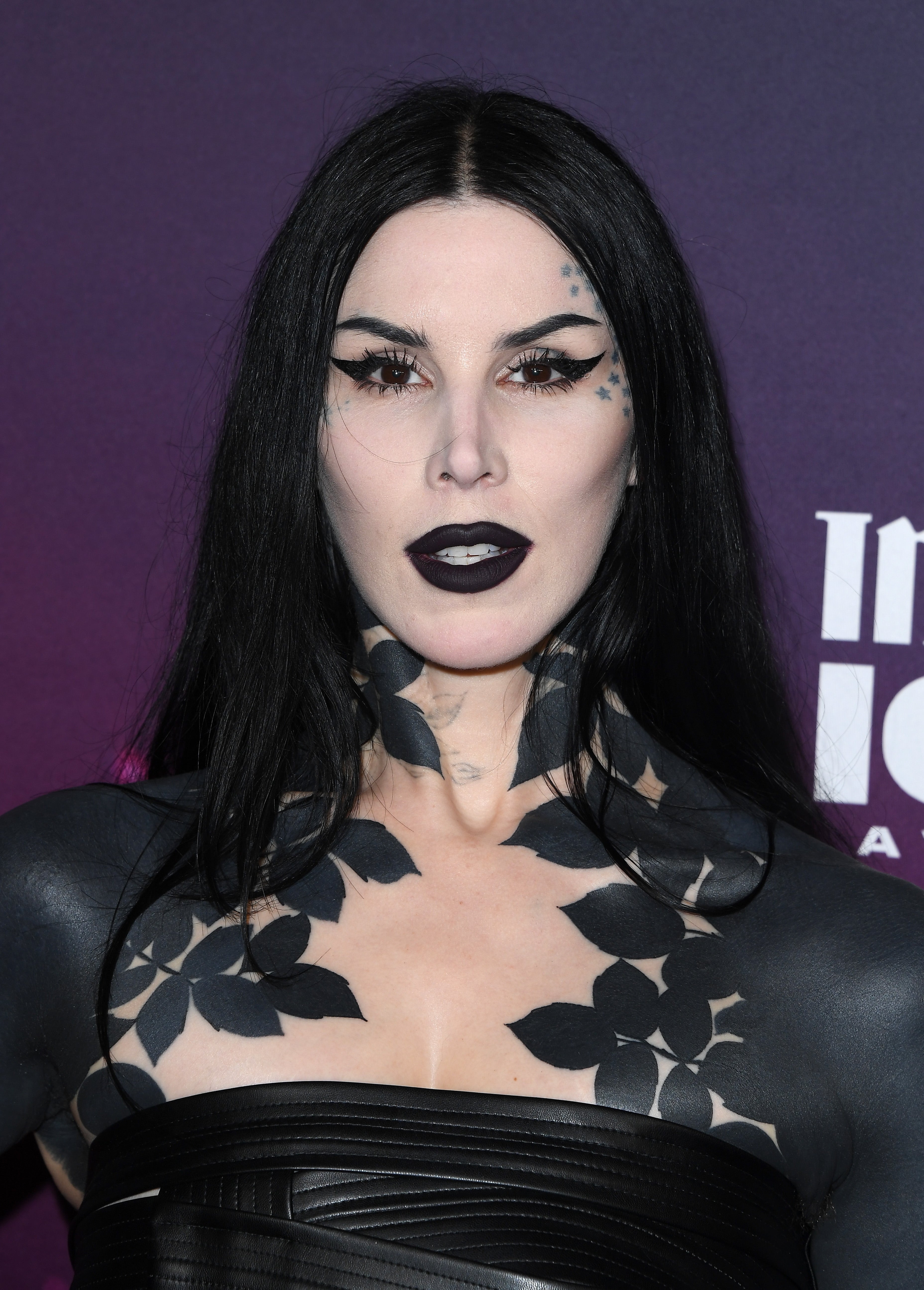 Person with agelong achromatic hair, bold eyeliner, and achromatic lipstick connected a reddish carpet. Wearing a strapless formal and intricate assemblage art